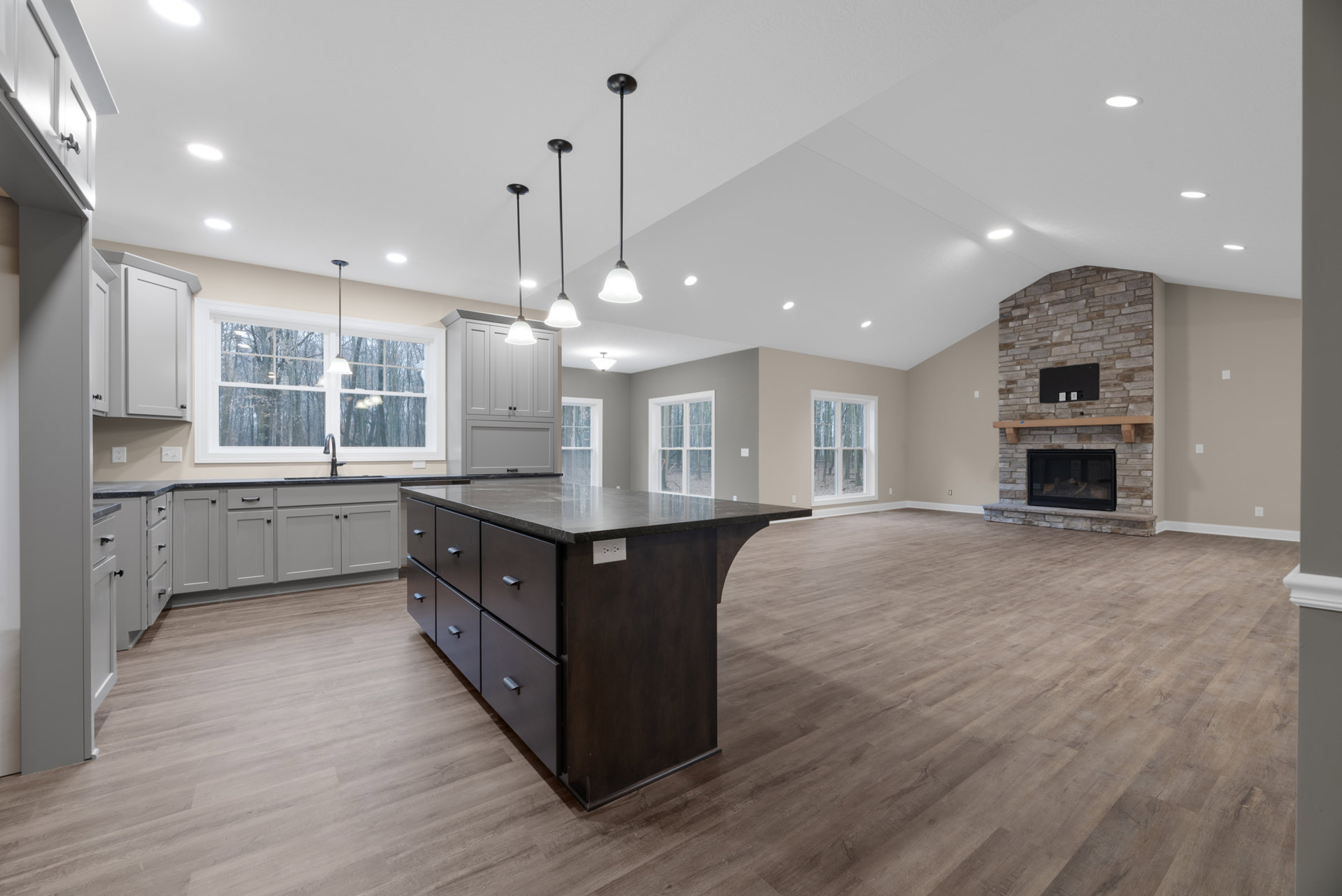 Spacious open plan kitchen featuring a central island with drawers, glass-door fireplace, large windows overlooking a forest, wood cabinetry, stone countertops, and visible