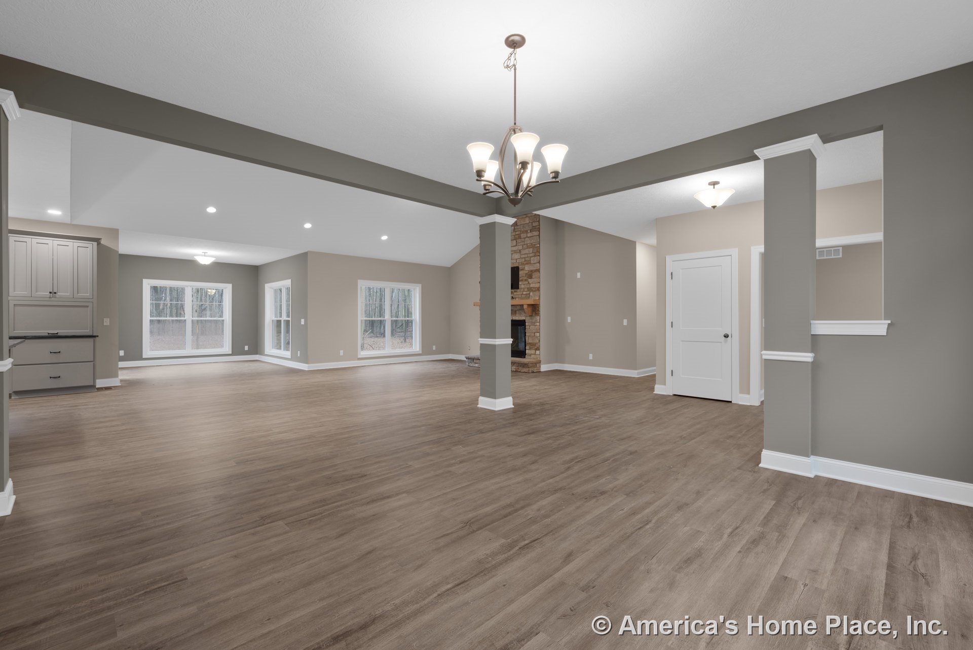Open living area with wood-look flooring, recessed ceiling lights, stone fireplace with wood mantel, built-in cabinetry with dark countertop, large windows framed in white trim