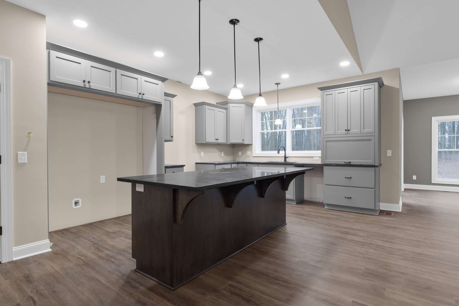 Spacious kitchen featuring a large marble-topped island, white cabinetry with black handles, pendant lights on black poles, wide window overlooking trees, and light wood flooring.