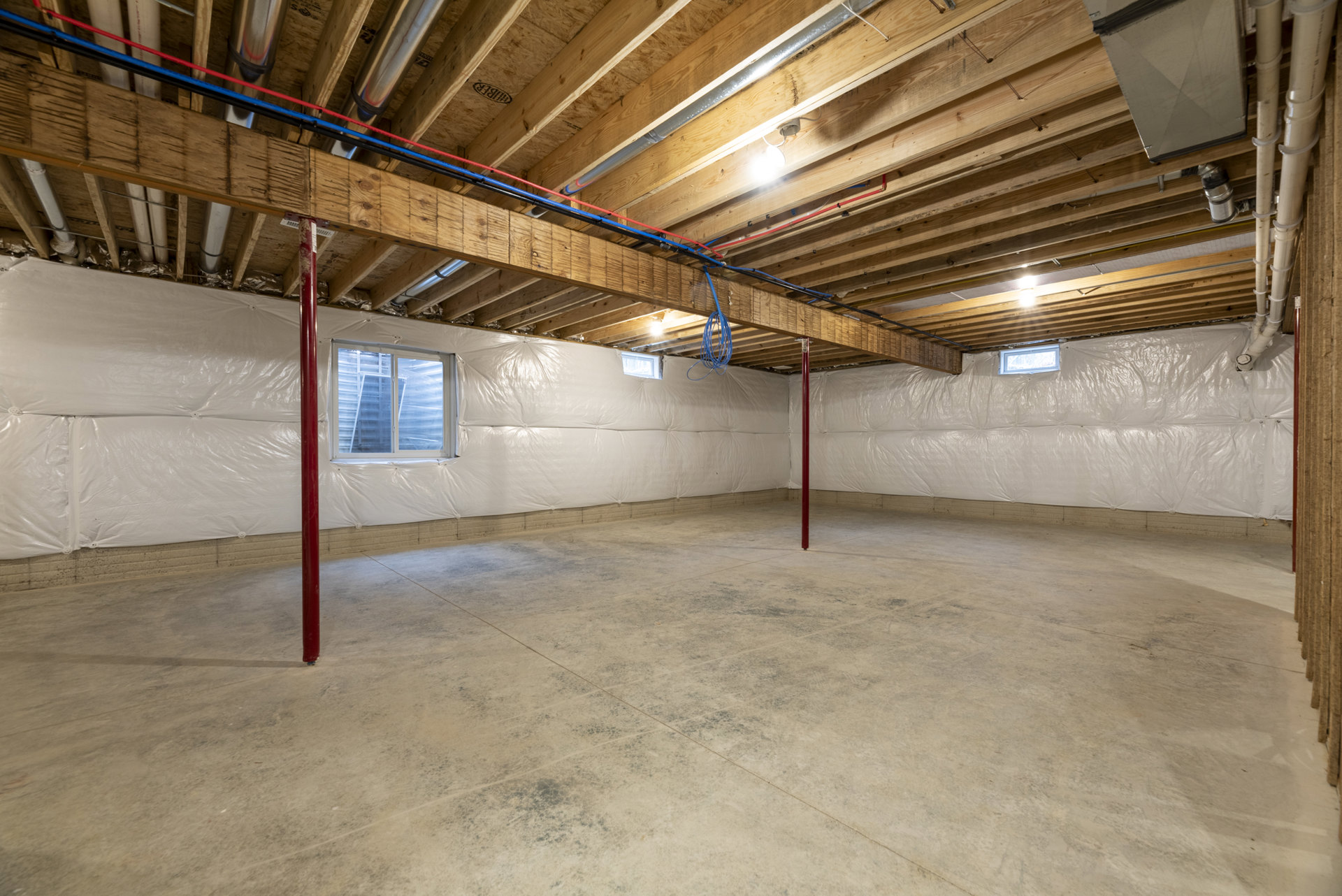 Room with exposed wooden ceiling beams, white-framed window, concrete floor, red support pole, visible pipes, and metal electrical box mounted on wood wall