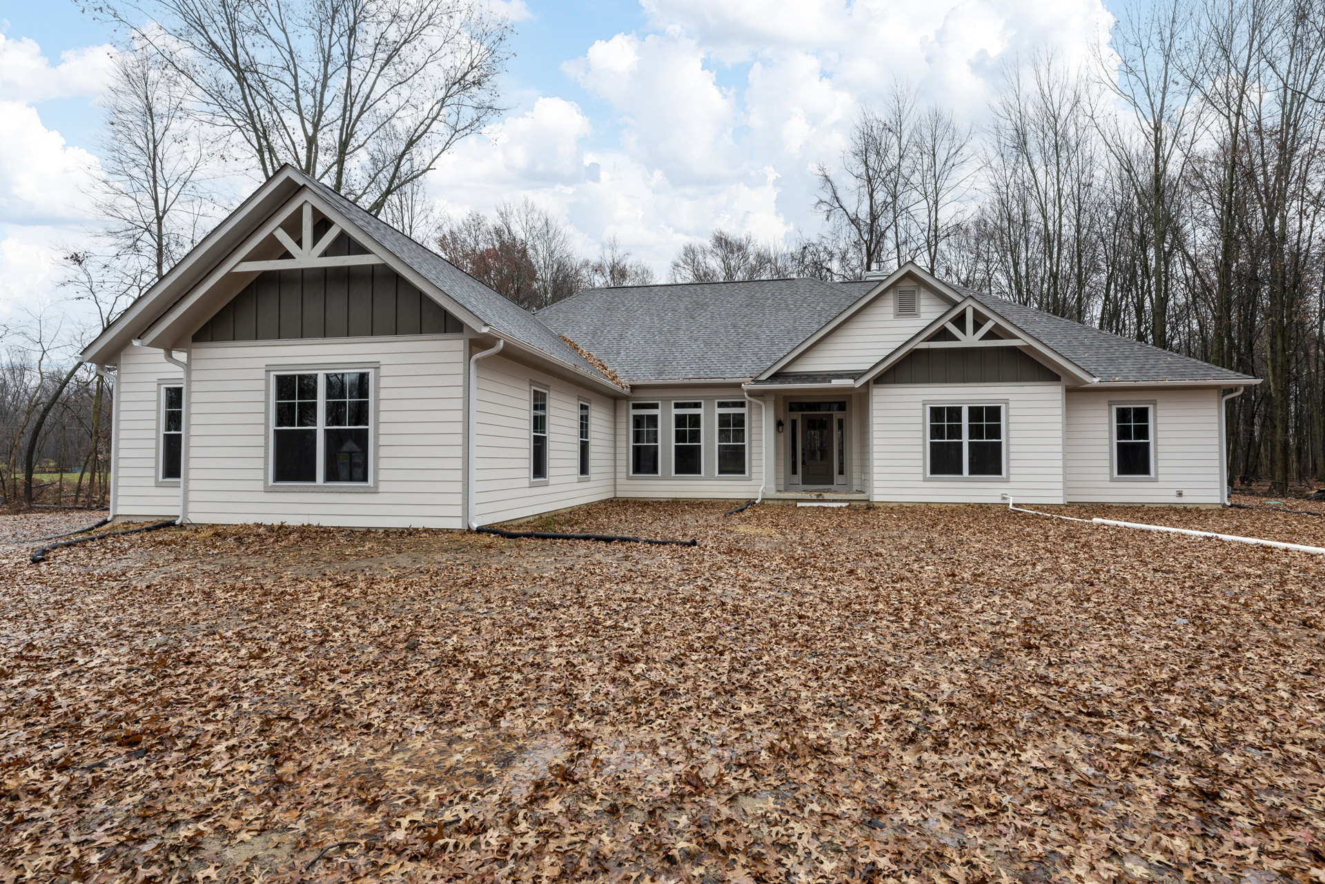 White-trimmed windows and front door on a cottage-style home, leaves scattered across the lawn, blue sky with clouds and mature trees in the background
