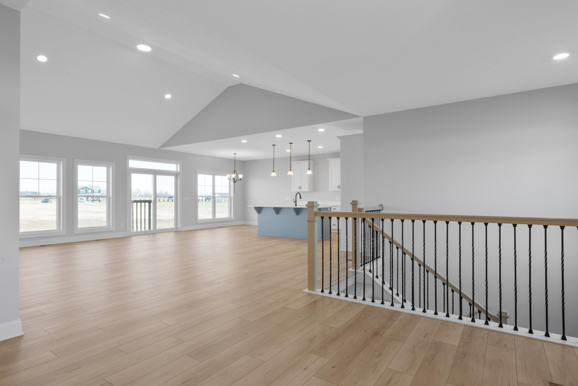 Spacious interior featuring wood flooring, modern staircase with metal railings, large windows revealing a blue house, car, fence, and grassy field outside