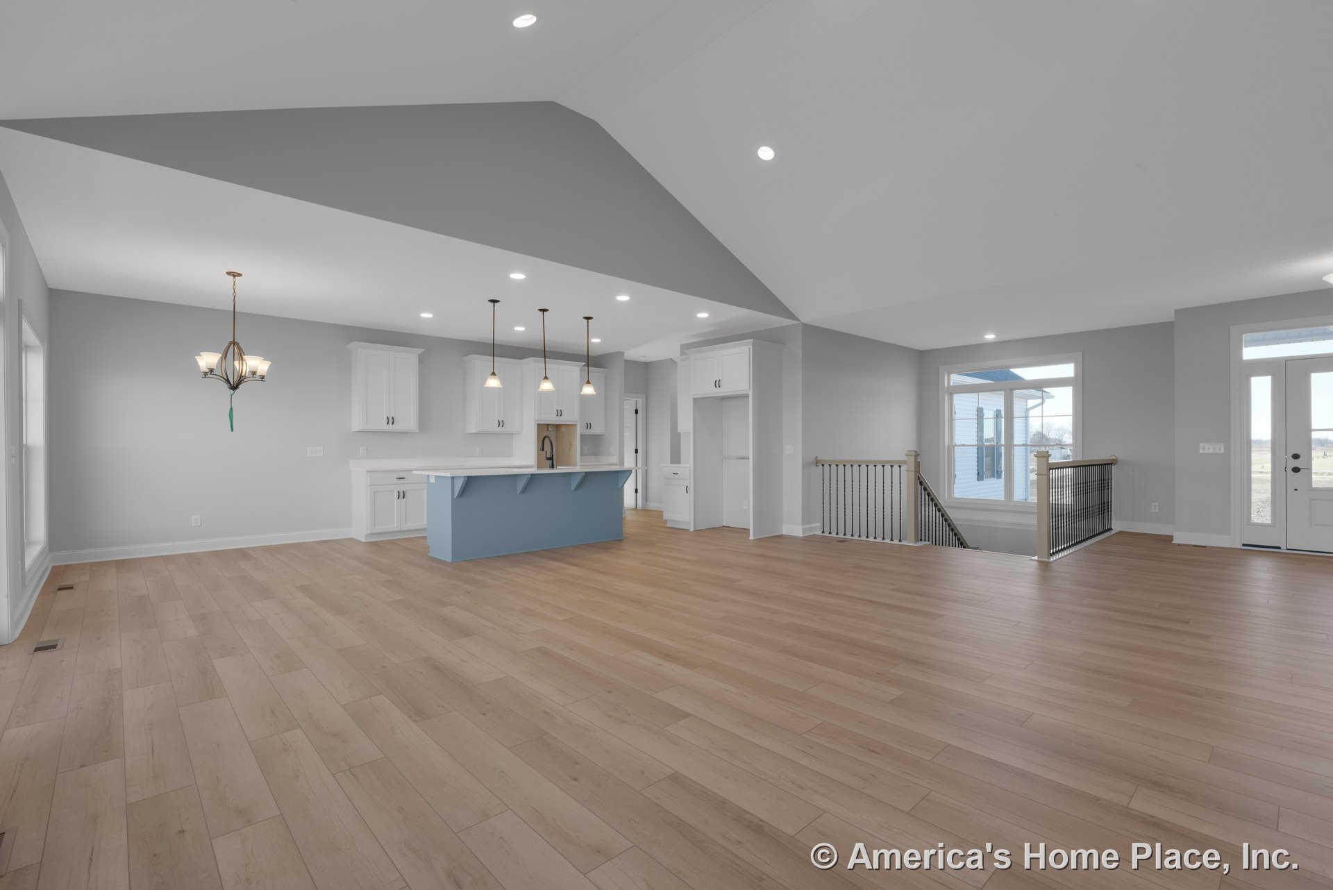 Vaulted ceiling and open floor plan with light wood plank flooring, white kitchen cabinets, blue kitchen island with pendant lighting, large windows and glass exterior door, stair