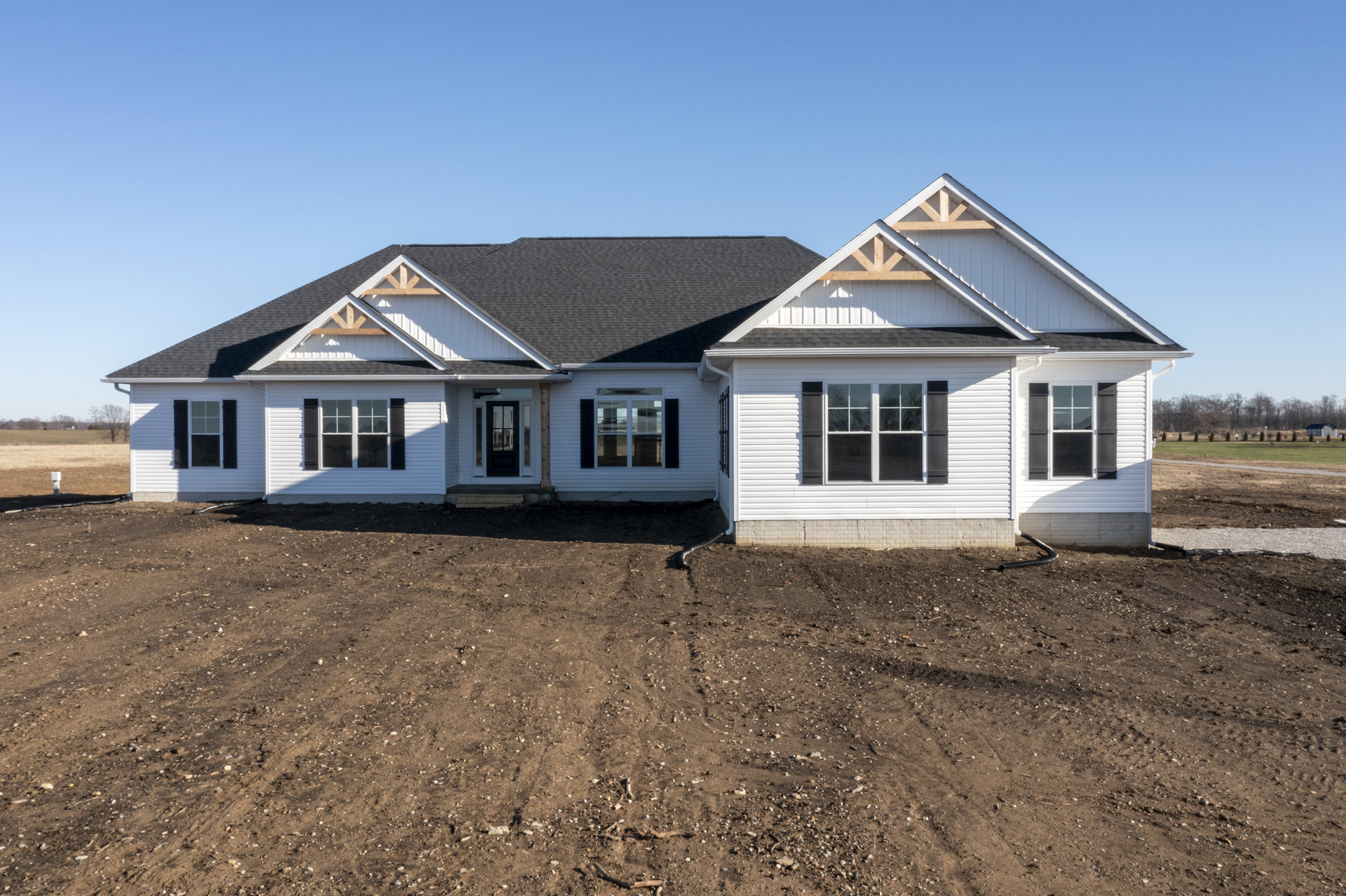 Modern house with black roof, light siding, multiple-pane windows, glass door, drain pipe, and unfinished dirt yard under clear sky