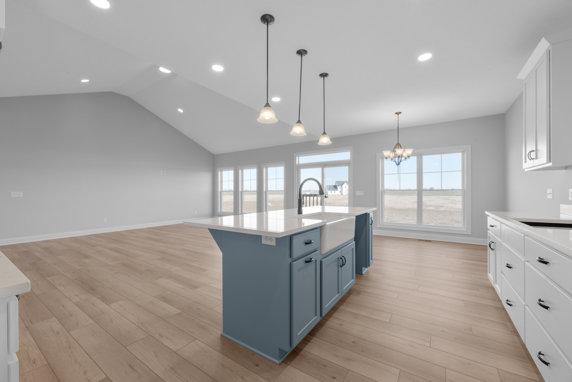 Kitchen with wood flooring, white cabinetry, central island featuring a built-in sink, pendant light fixtures, large window overlooking a grassy field, and stainless steel accents