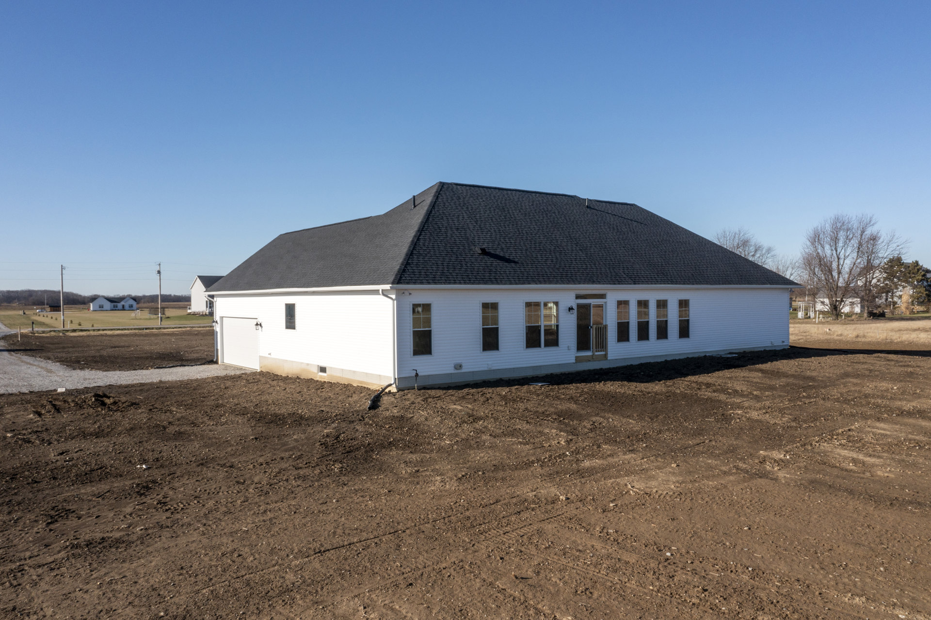 White siding house with black shingle roof, large windows, front door with exterior light, surrounded by dirt lot and scattered trees, black pipe lying on ground.