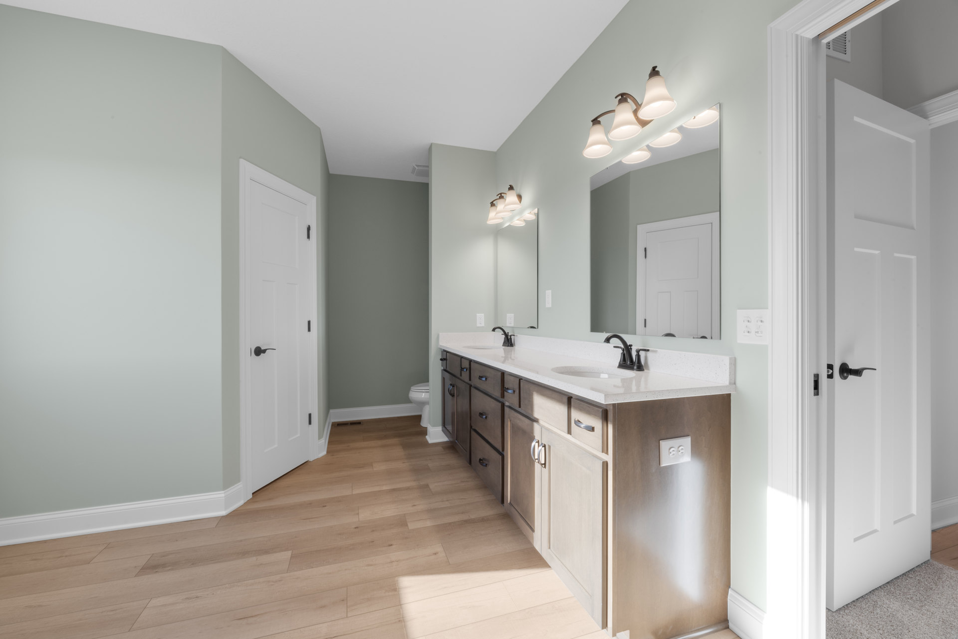 Bathroom with double white sinks, quartz countertop, large framed mirror, row of modern light fixtures above, white cabinetry, tile flooring, and a white door with black handle