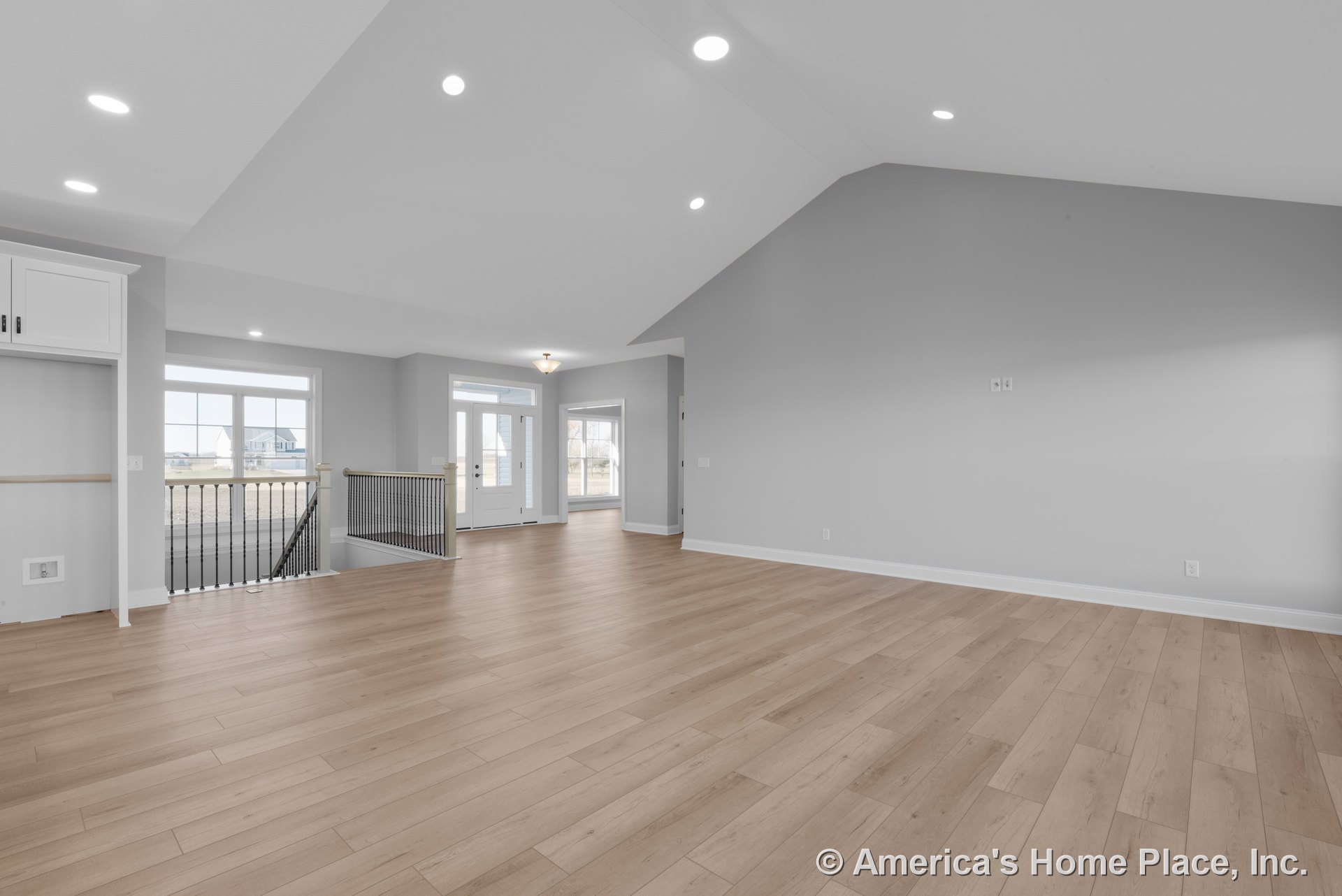 Open living area with light wood plank flooring, vaulted ceiling with recessed lighting, expansive windows framed in white trim, open stair railing featuring black balusters, white