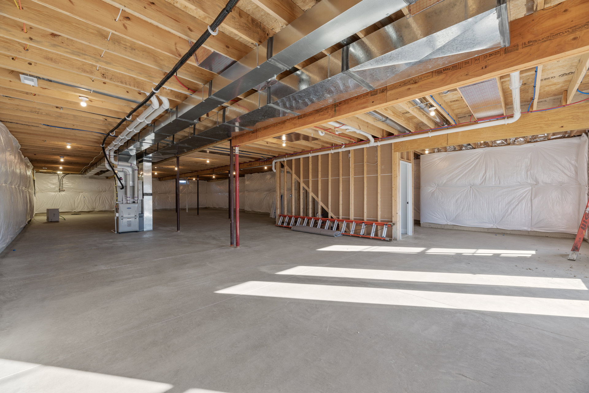 Spacious unfinished room with exposed metal ceiling beams and pipes, white plastic wall covering, rolled carpet on floor, and ladder against wall