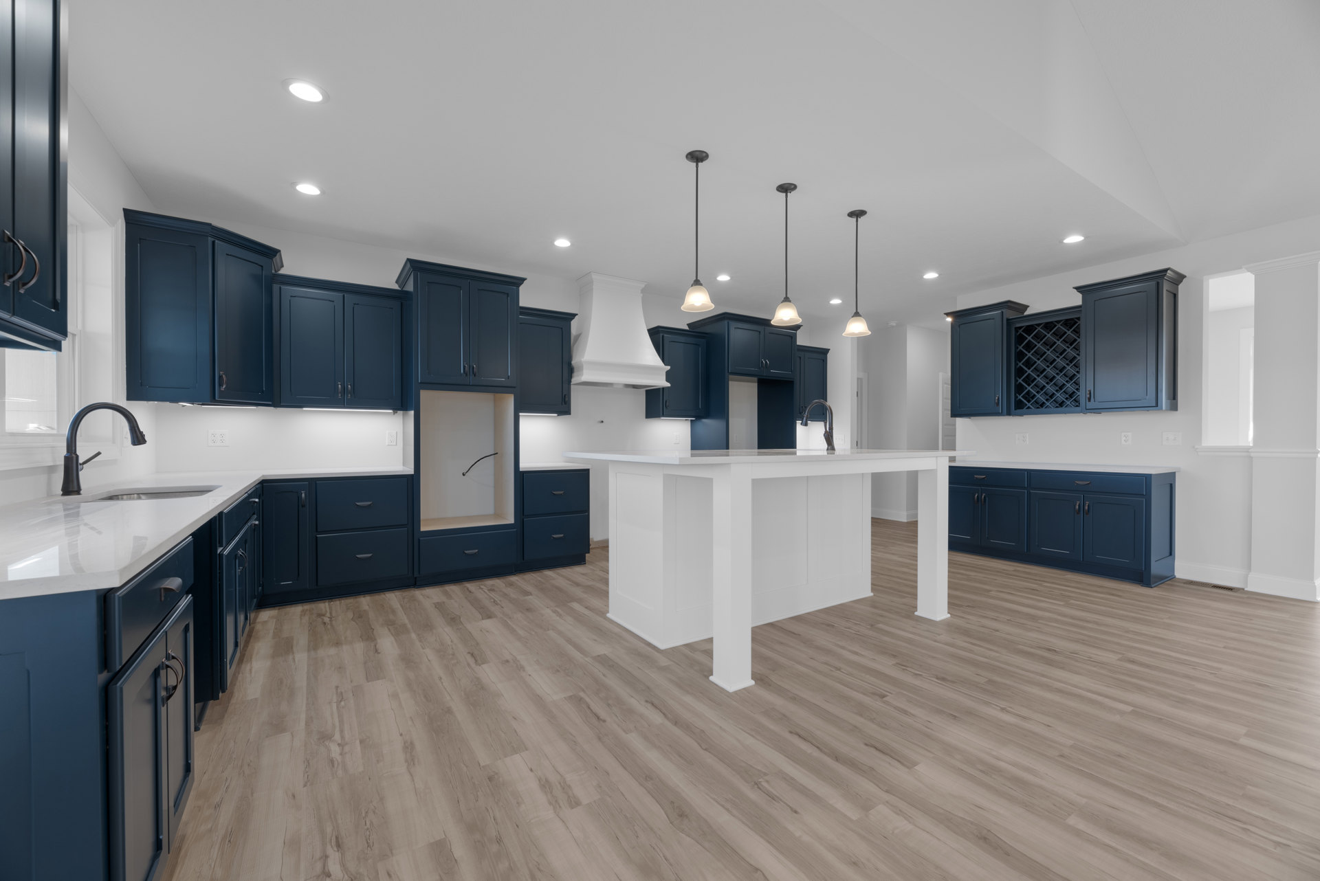 Blue shaker cabinets with brushed metal hardware, white quartz countertops, wide-plank wood flooring, central white kitchen island, stainless steel sink, and large window with