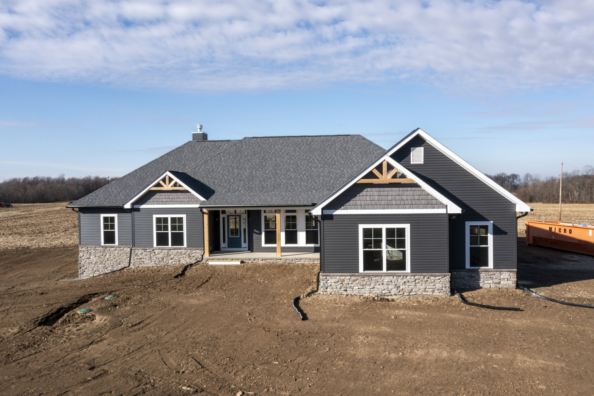 Two-story house under construction with exposed framing, white window frames, covered porch, black pipe protruding from siding, dirt hill in foreground, cloudy sky overhead
