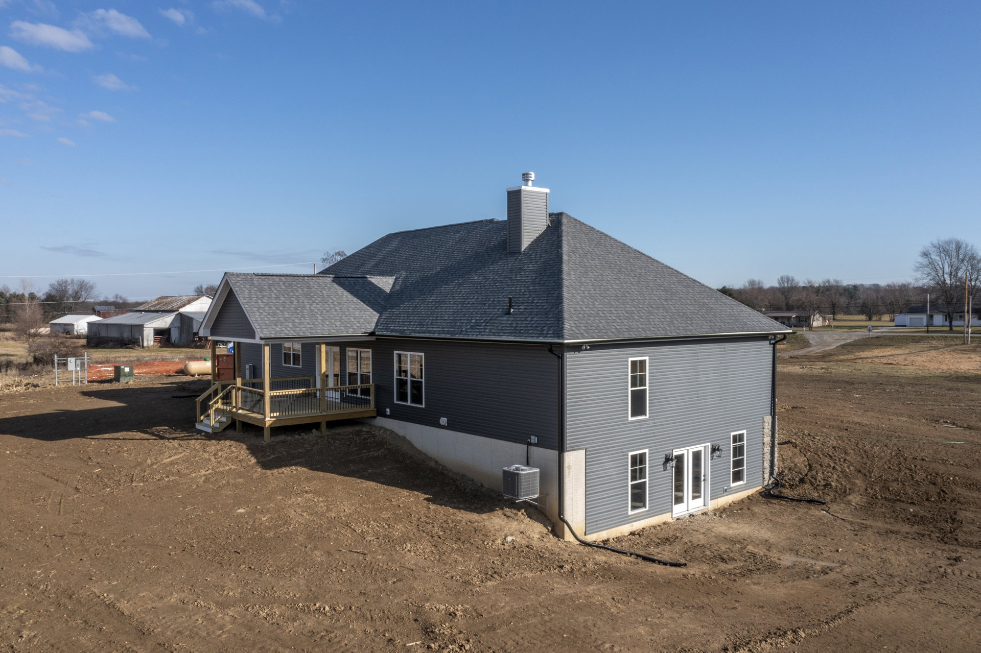 Two-story house with gray siding, white trim, wooden deck featuring metal railing, covered porch, multi-pane windows, roof with brick chimney, large air conditioner unit on deck