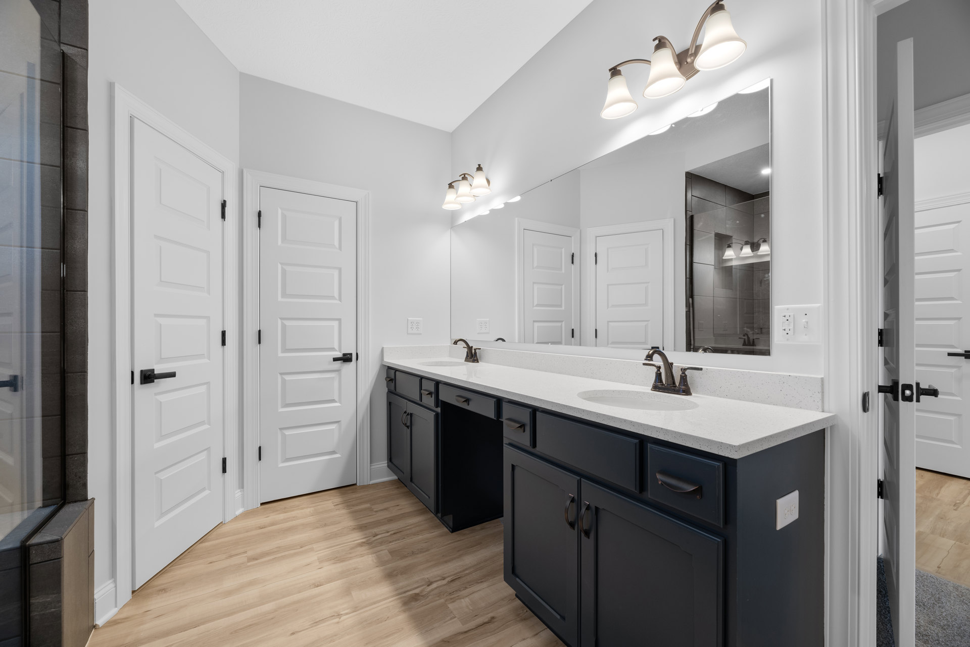 Bathroom with double sinks and wide mirror, white cabinetry, black hardware, wood flooring, three-light fixture above mirror, white door with black handle