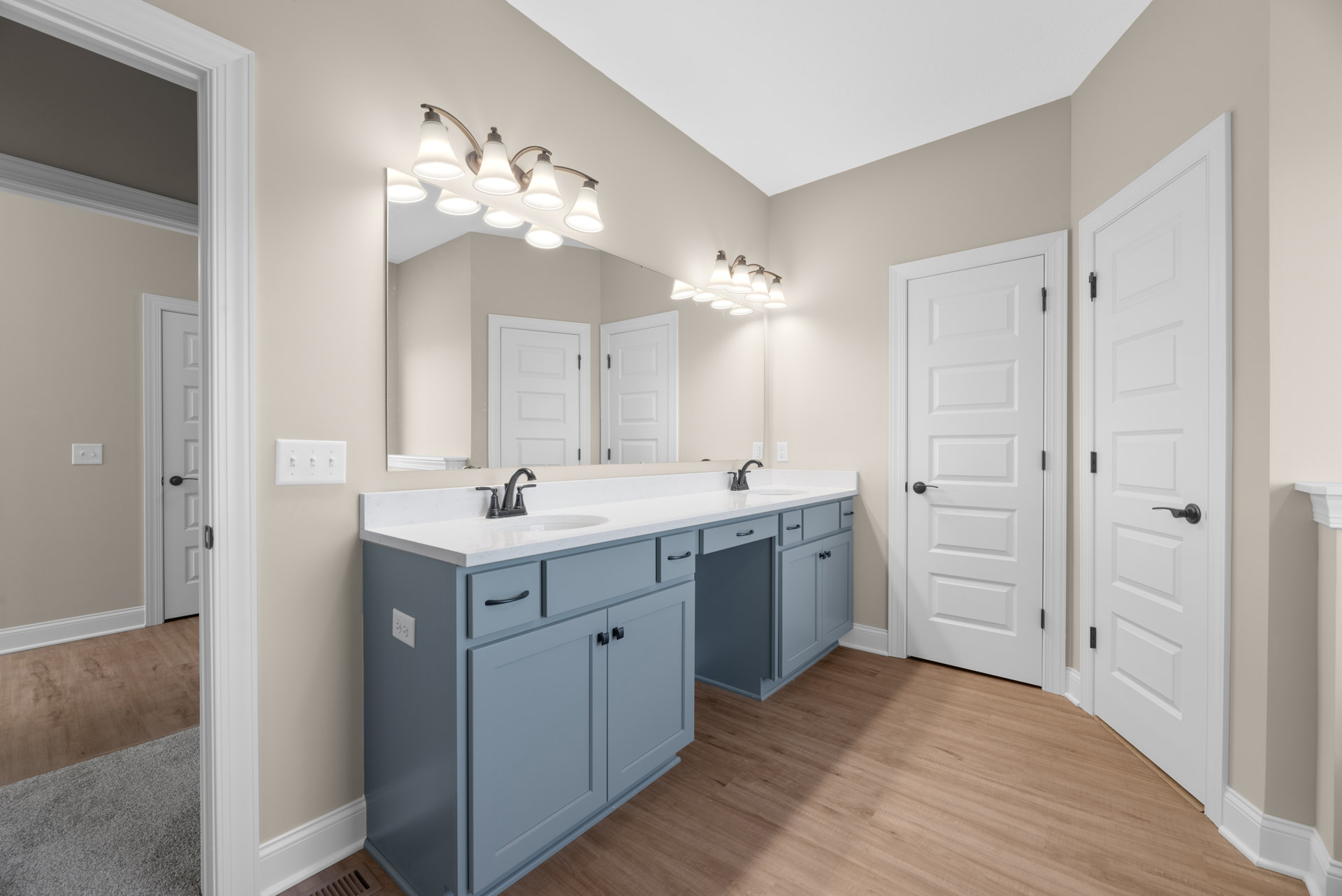 Bathroom with white double vanity, quartz countertop, rectangular mirror, row of modern light fixtures above, white door with black hardware, light gray tile flooring, shaker-style