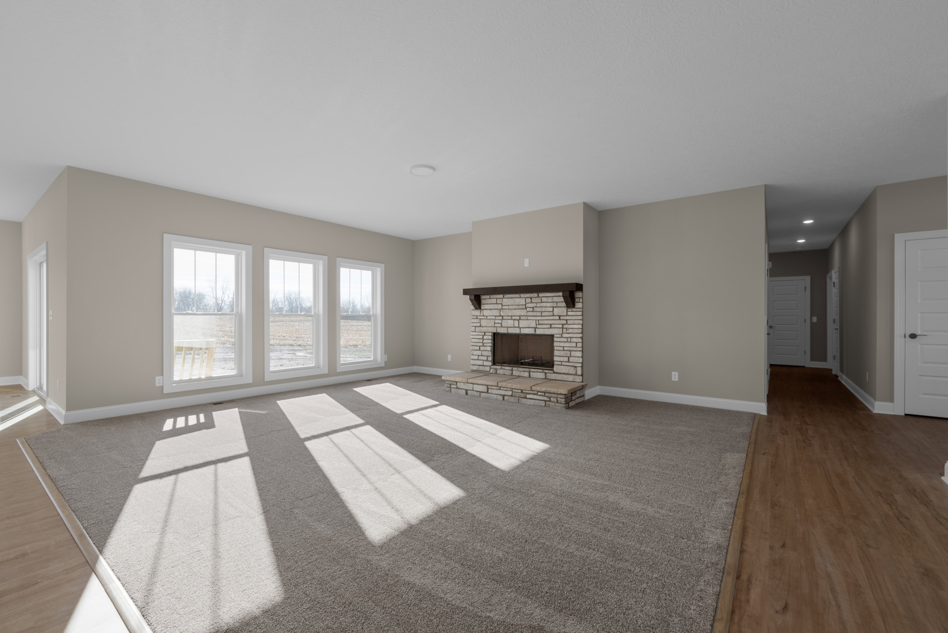 Living room with plush carpet flooring, white plaster walls, central stone fireplace, large windows overlooking grassy field, wood trim, and recessed ceiling lighting
