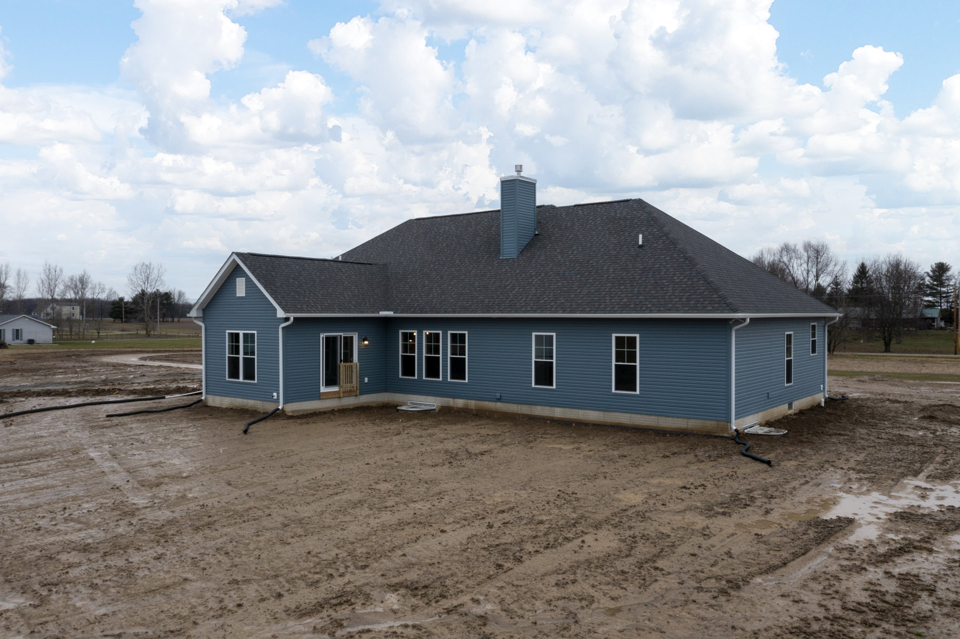 Partially built house with exposed framing, chimney on shingled roof, muddy ground in foreground, wooden fence beside entry door, blue building visible in background under cloudy
