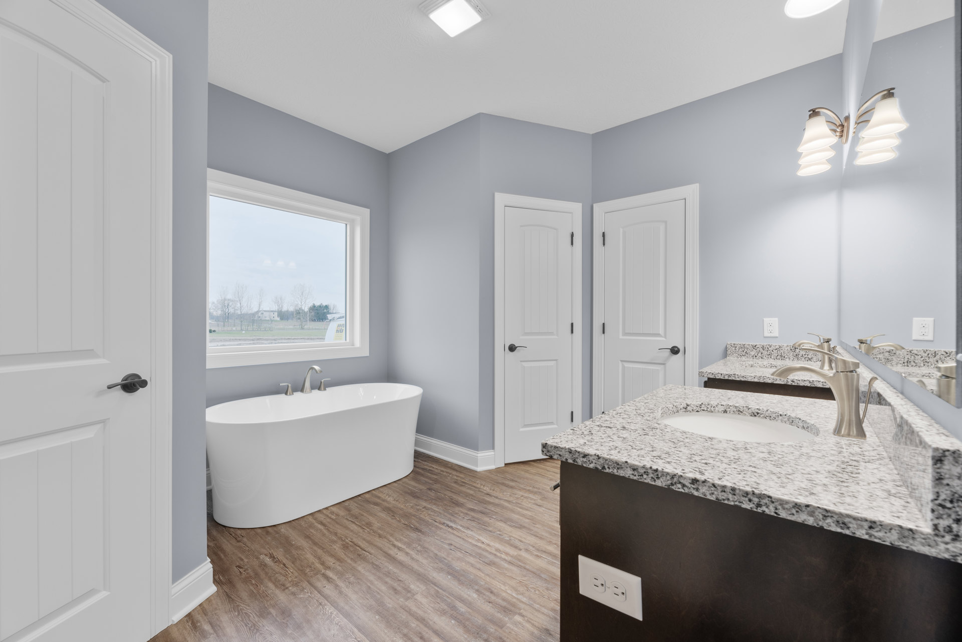 White freestanding bathtub with silver faucets beside a window overlooking trees and grass, light fixture above, bathroom sink with mirror and chrome faucet, tiled walls, and door