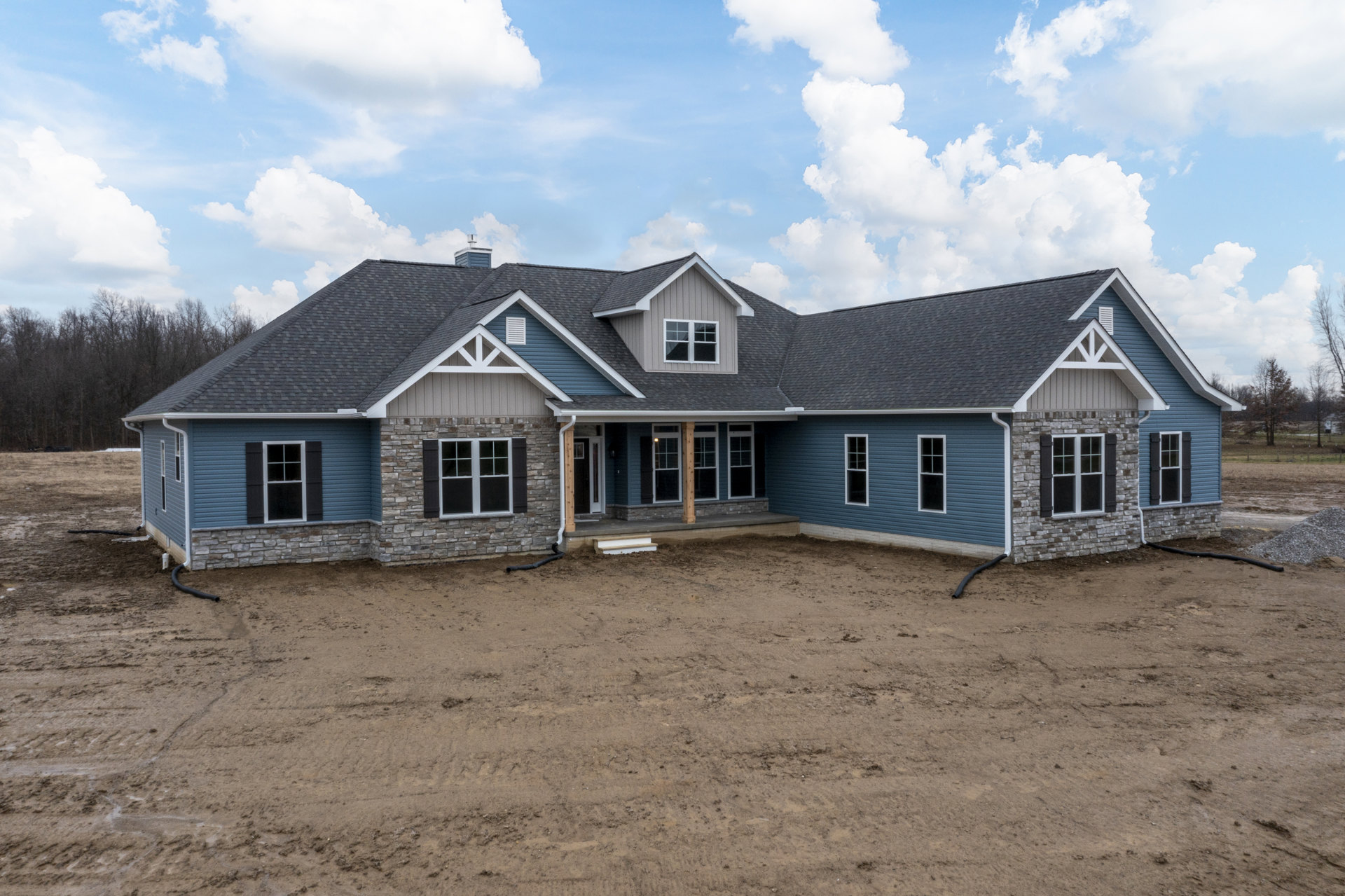 Two-story house with white-trimmed windows, beige siding, and a dirt yard with exposed pipe under a partly cloudy blue sky