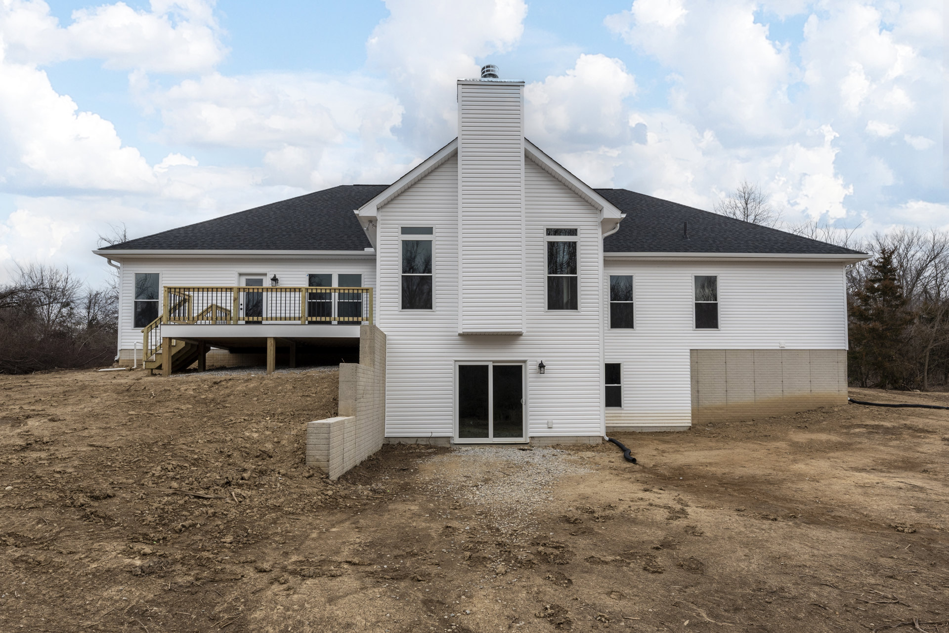 White cottage-style home with a brick chimney, sliding glass door opening onto a wooden deck with railing, front porch, and windows reflecting nearby trees.