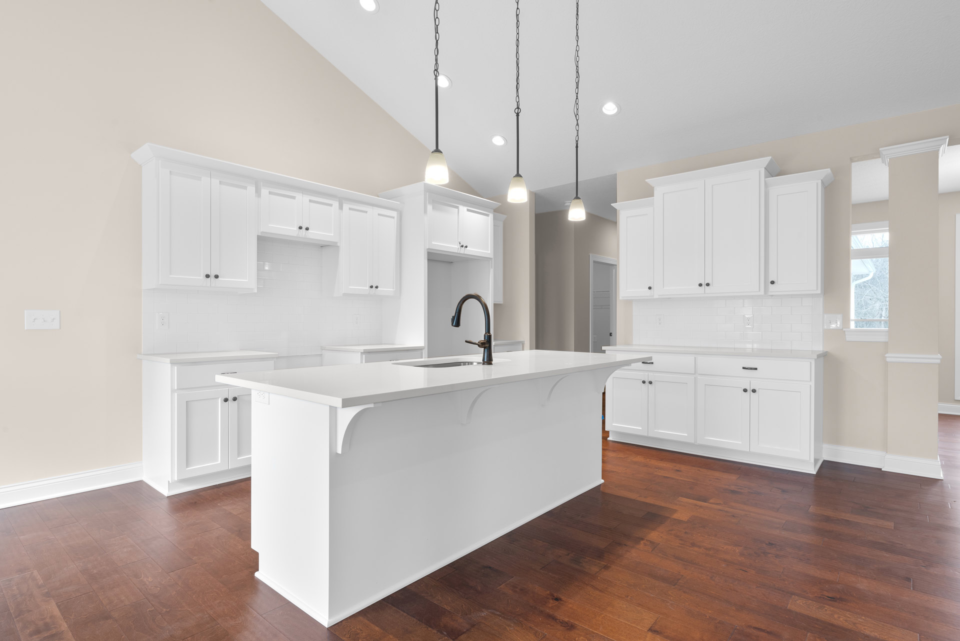 White kitchen with large central island, white cabinetry, wood flooring, stainless steel faucet, pendant lights, and window with white trim.