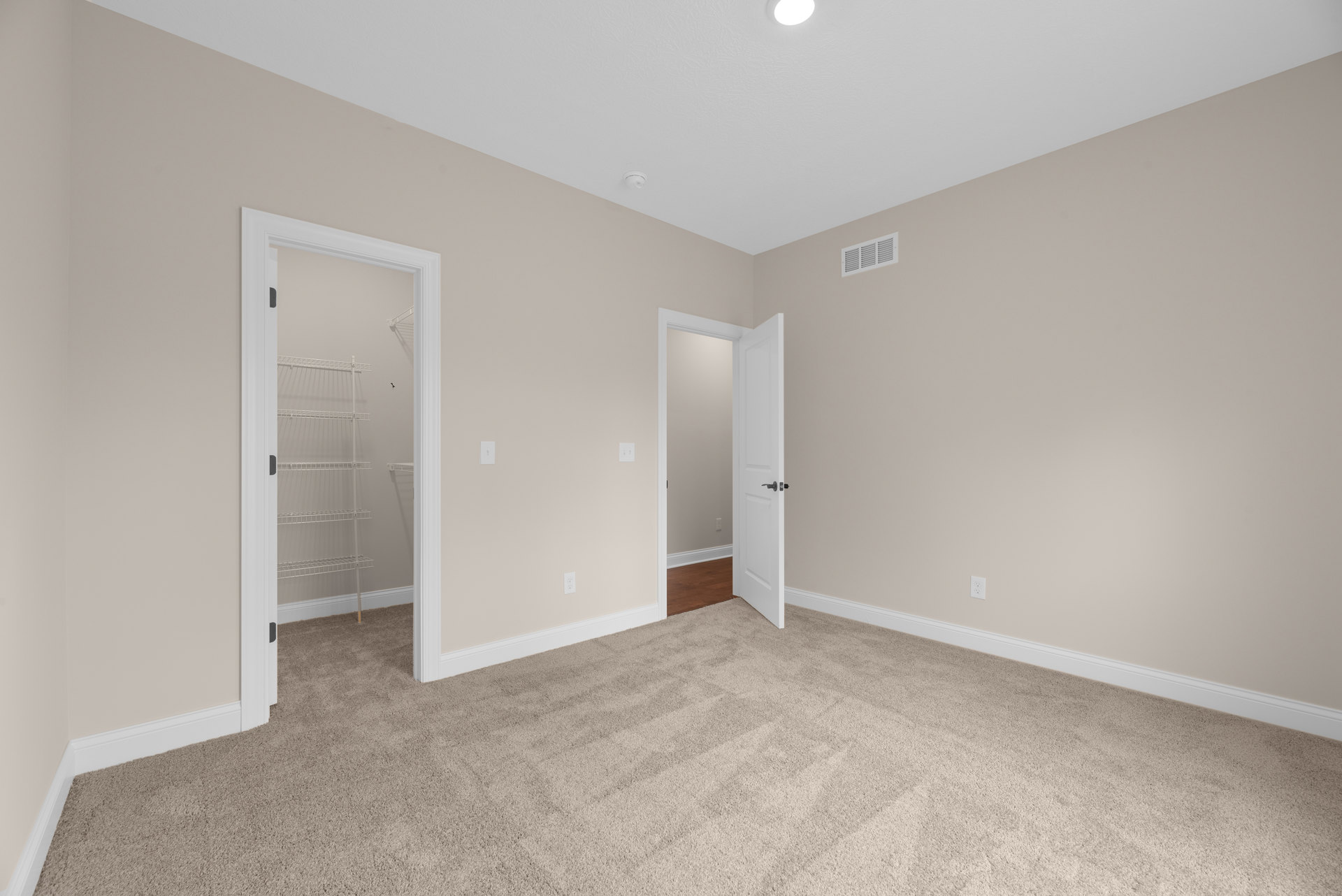 Beige-walled room with laminate flooring, open white door featuring a black handle, white vent on wall, and white metal shelf in adjacent space