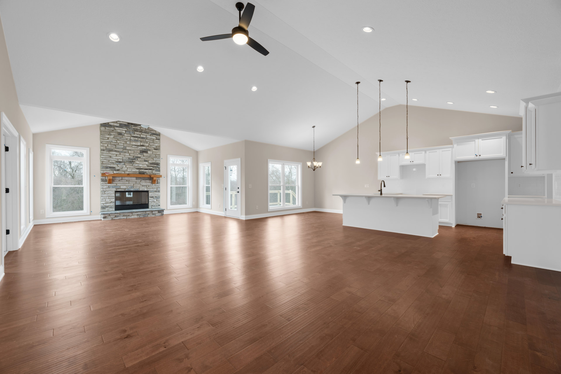 Spacious living area with hardwood floors, stone fireplace, built-in bar, ceiling fan with light, large windows overlooking deck and trees