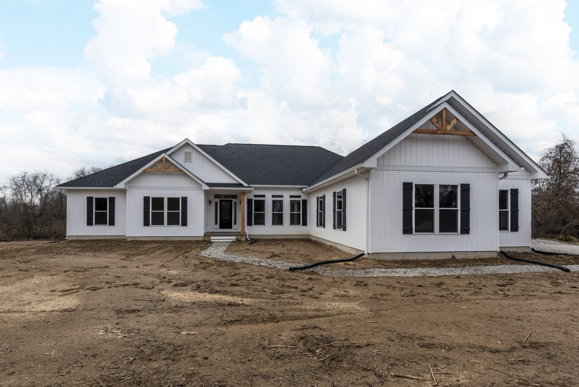 White house under construction with black roof, black shuttered windows, exposed dirt yard, PVC pipe, and cloudy sky