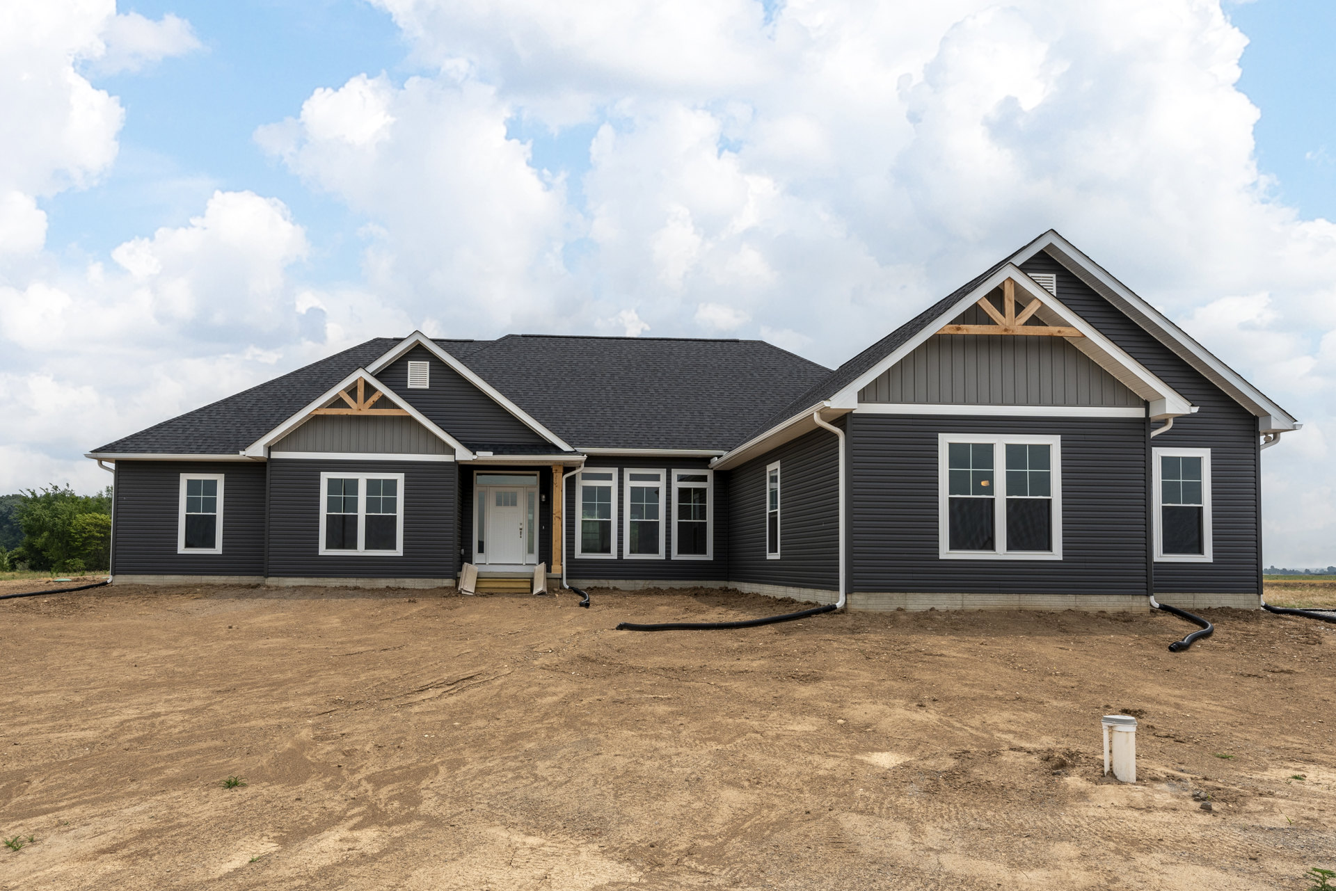Partially built house with exposed framing, white-trimmed windows, white pipe in dirt foreground, covered porch, shingled roof, and cloudy blue sky overhead
