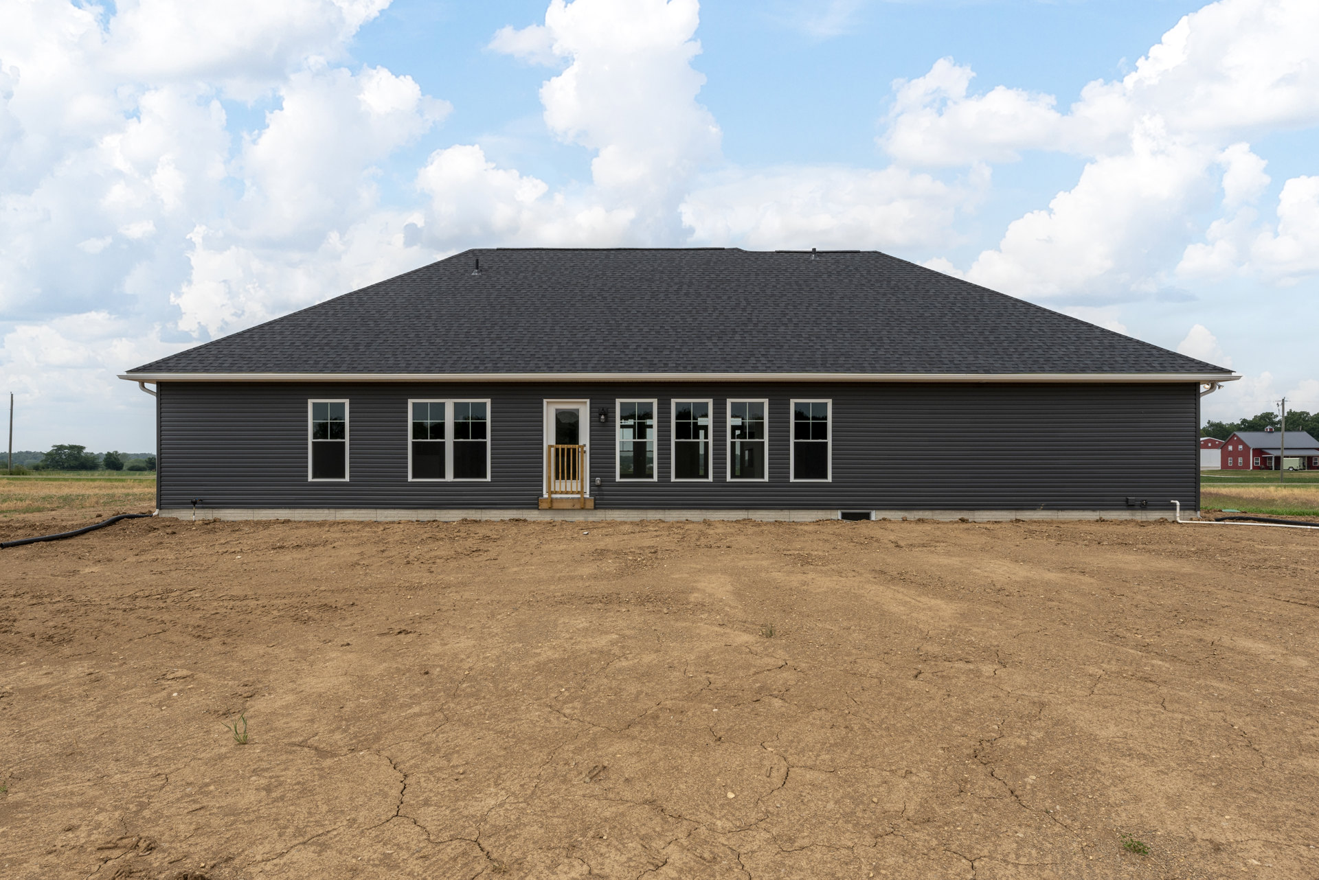 Single-story house with white-framed windows, wooden porch railing, and a front door, set against a large dirt field under a blue sky with scattered clouds
