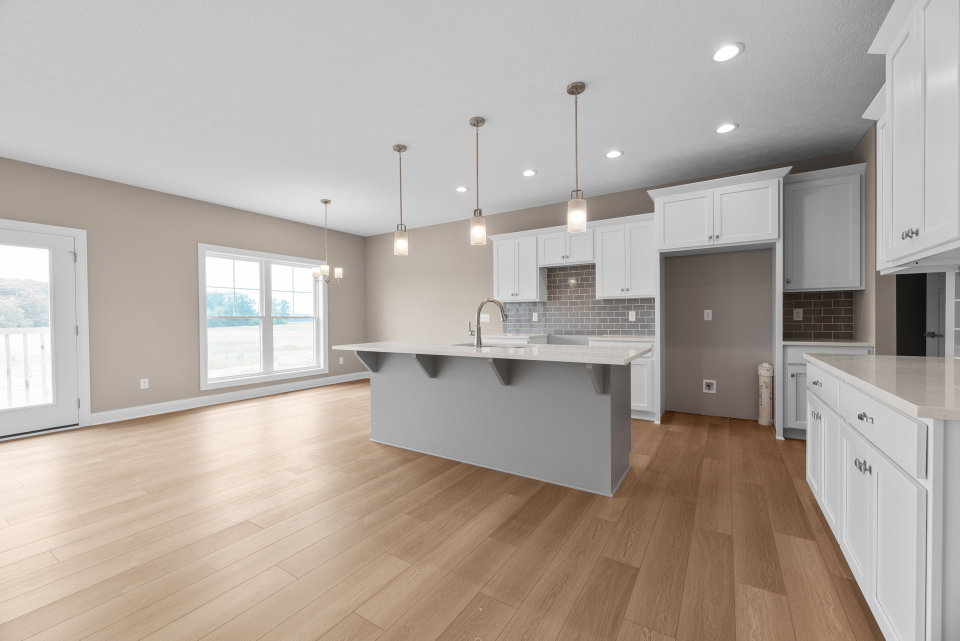 Kitchen with white island countertop and built-in sink, wood laminate flooring, white cabinetry, large window overlooking grassy field, white door with railing and trees visible