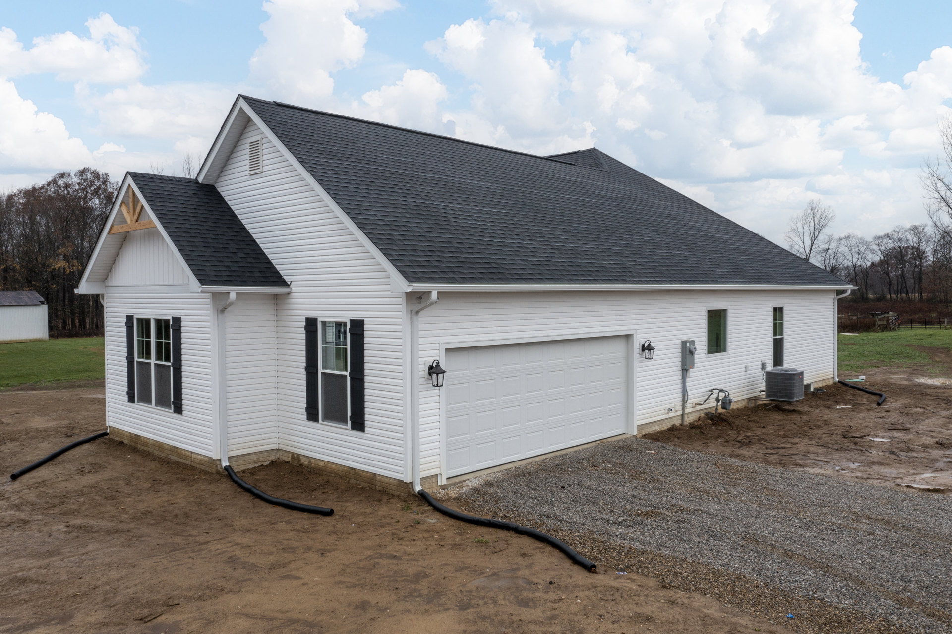 White house with black roof and black shuttered window, attached garage, large grey heat pump beside exterior wall, cloudy sky overhead, tree in background