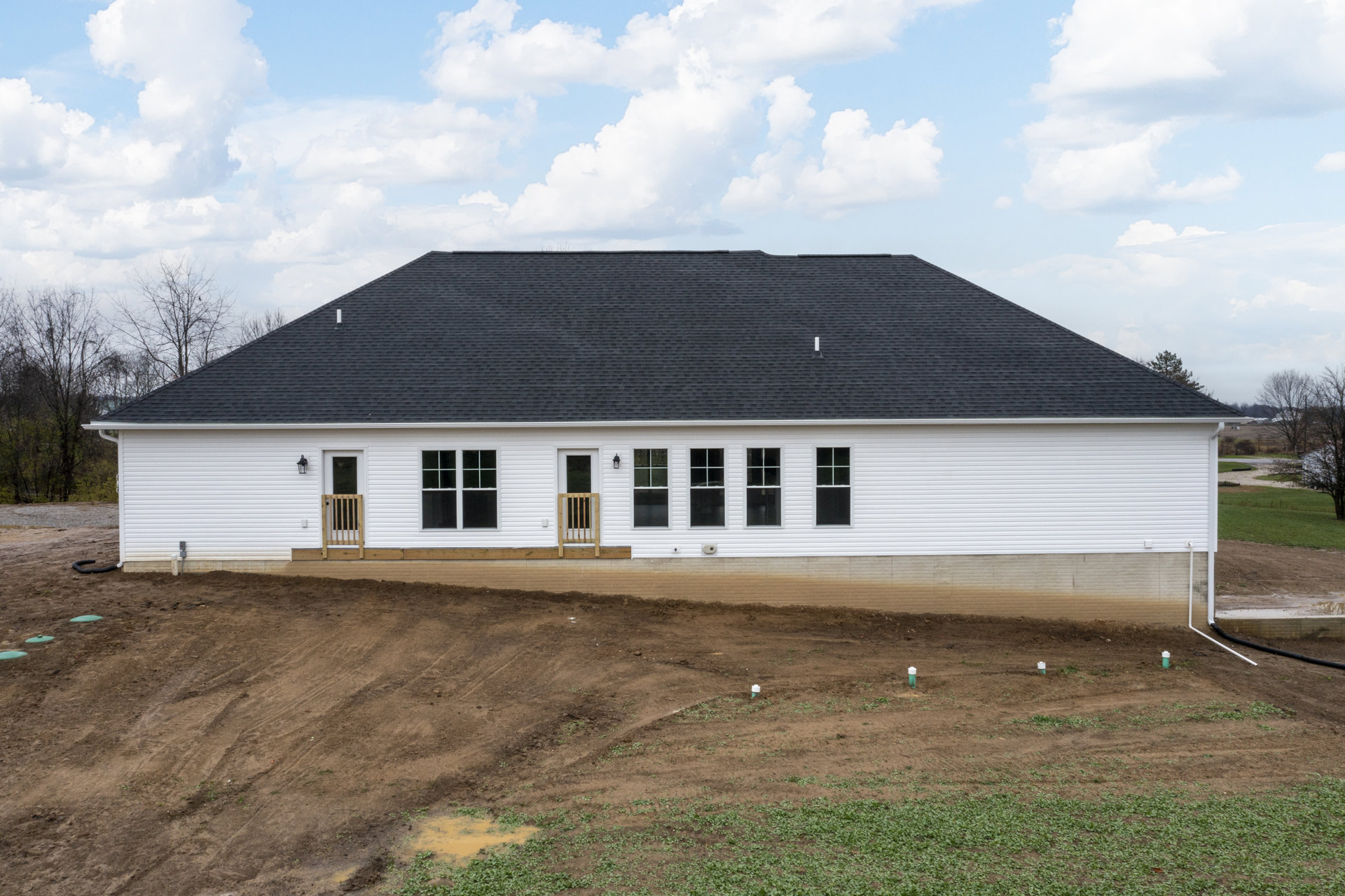 White siding house with black shingle roof, wooden window frame, white-framed window, dirt yard, wooden gate set in white wall, grassy lot, trees in background, cloudy sky overhead