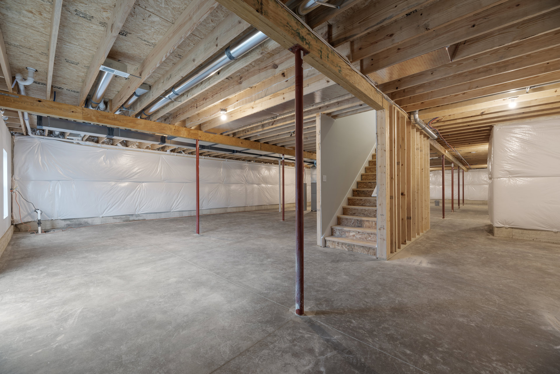 Basement room with exposed wooden ceiling beams, metal pipe, concrete floor, red support pole, and unfinished staircase leading upward.