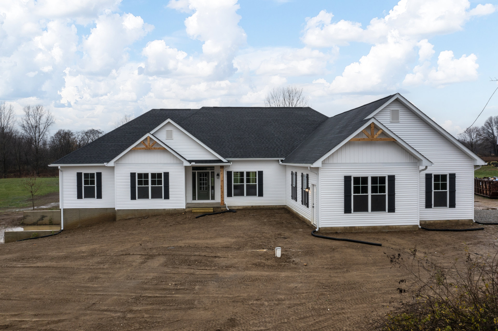 Modern house with light-colored siding, multiple windows, and a dirt yard; tree branches frame the roofline under a cloudy sky.