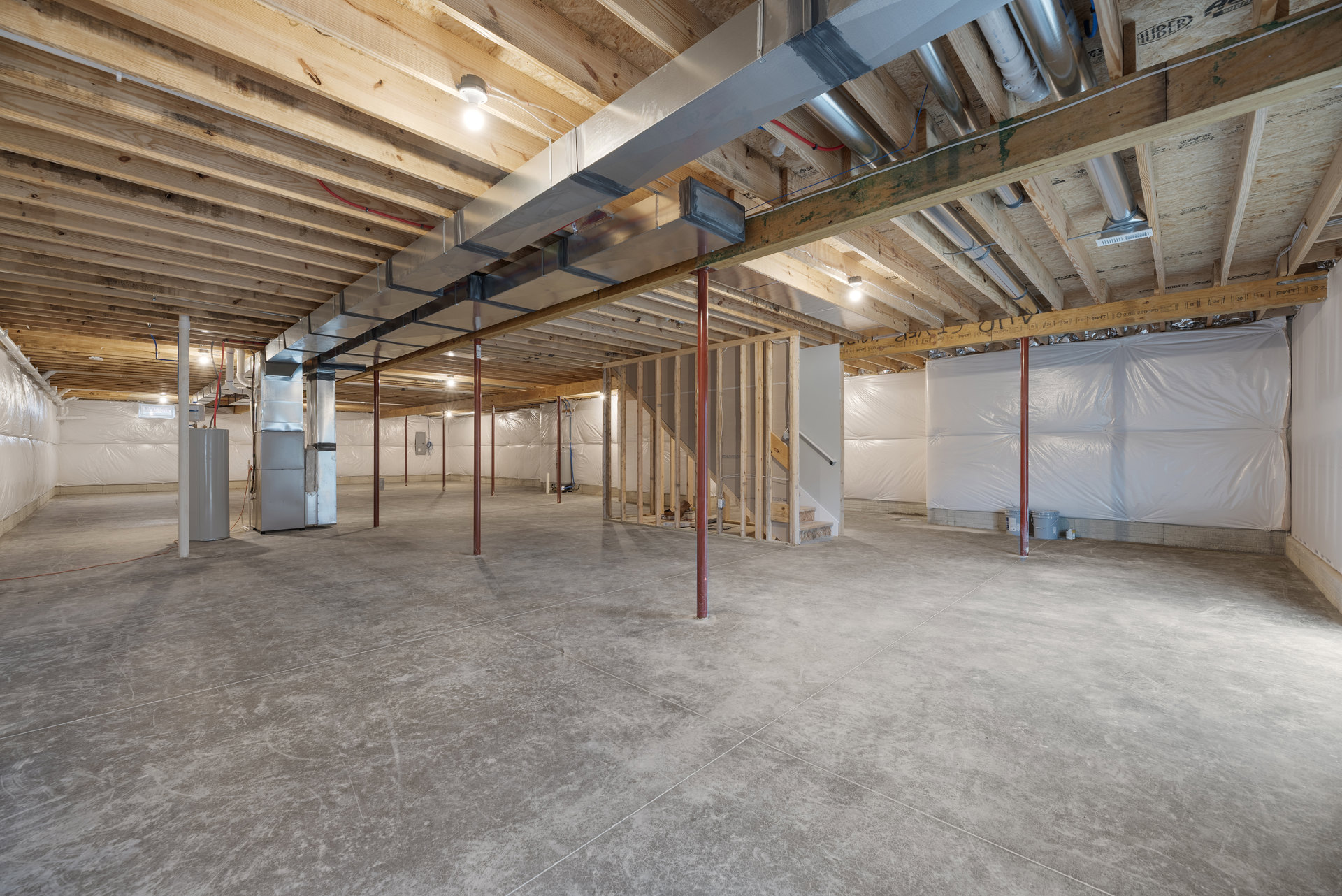 Exposed metal beams and pipes on ceiling, concrete floor with red support pole, white pillar with black handle, single light bulb, unfinished basement interior