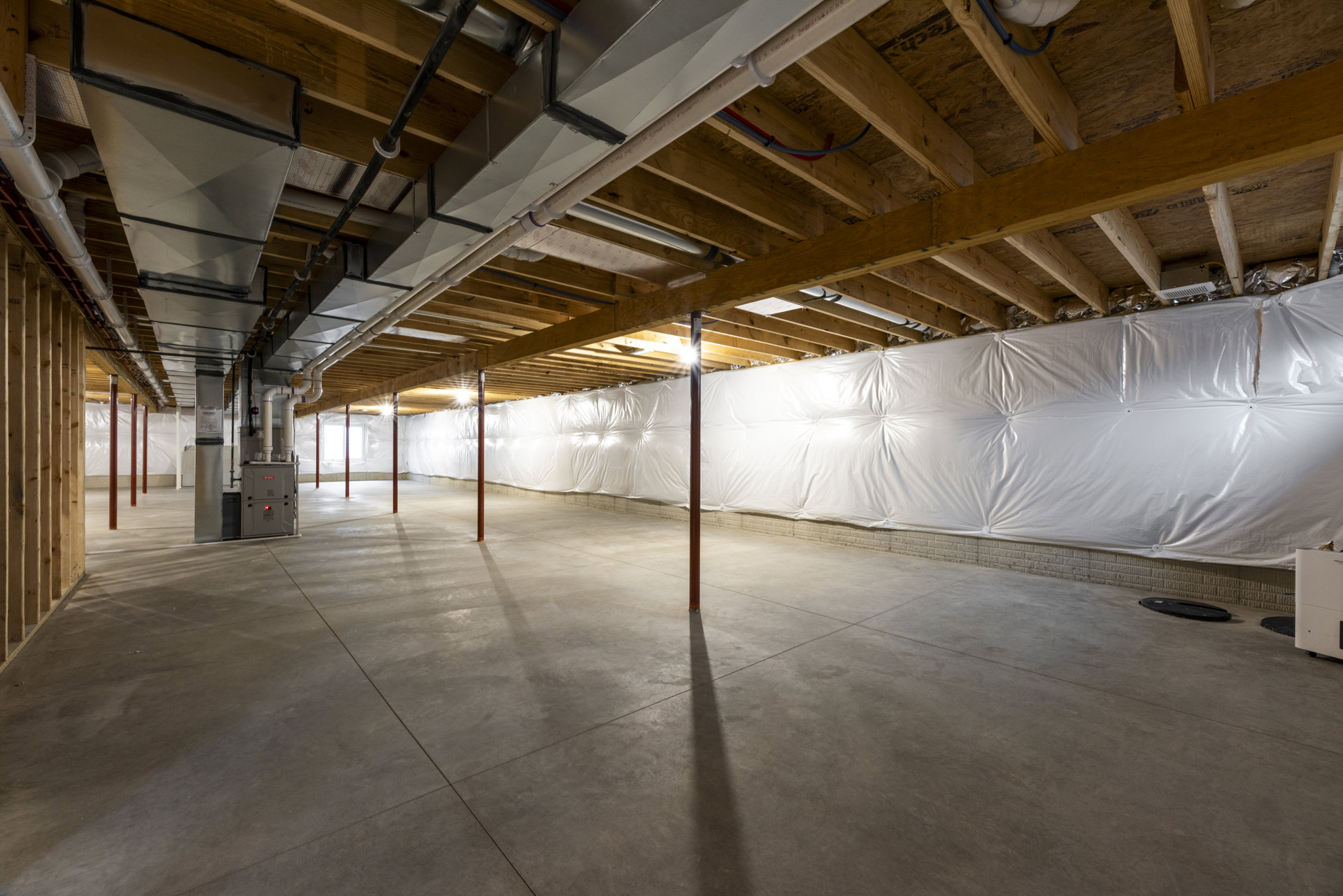White wall with exposed wooden ceiling beams, visible pipes and lighting fixtures overhead, concrete floor partially covered with white plastic sheeting, empty interior space.