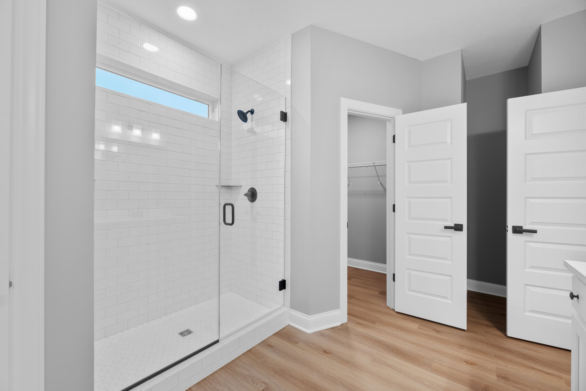 Bathroom featuring a frameless glass shower enclosure, wood plank flooring, tile shower wall with chrome shower head, recessed ceiling light, white door with black handle, and