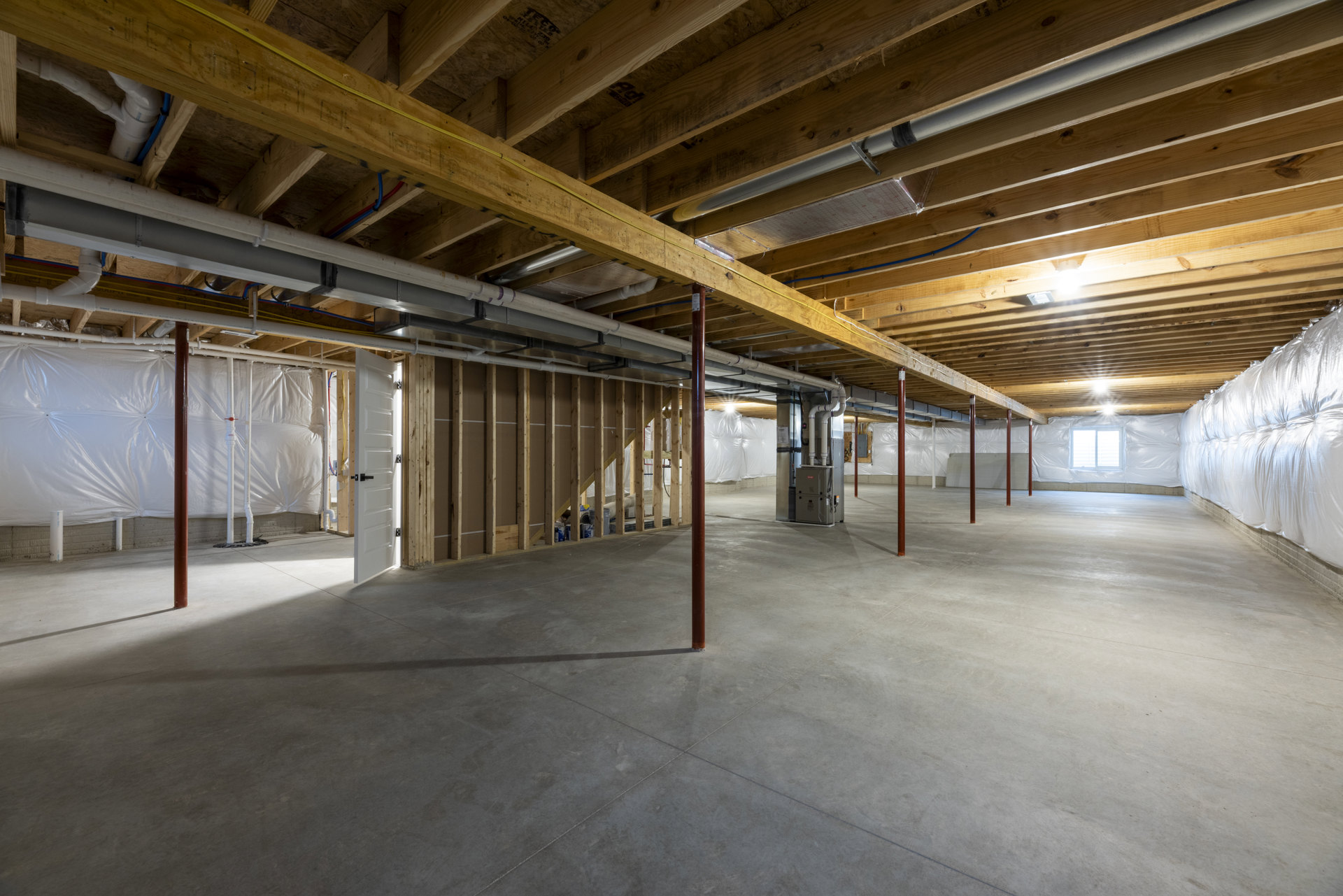 Spacious basement with exposed wood beam ceiling, visible metal pipes, concrete floor, metal pole, large white bag against wall, and metal box.
