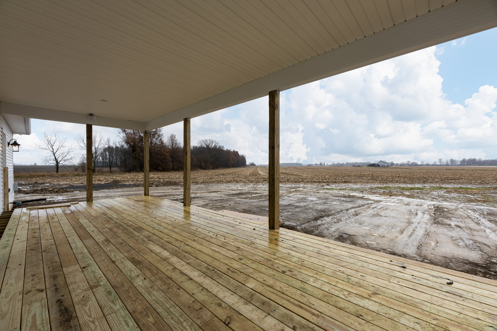 Wooden deck with sturdy pillars overlooking a dirt field, trees and cloudy sky in the background