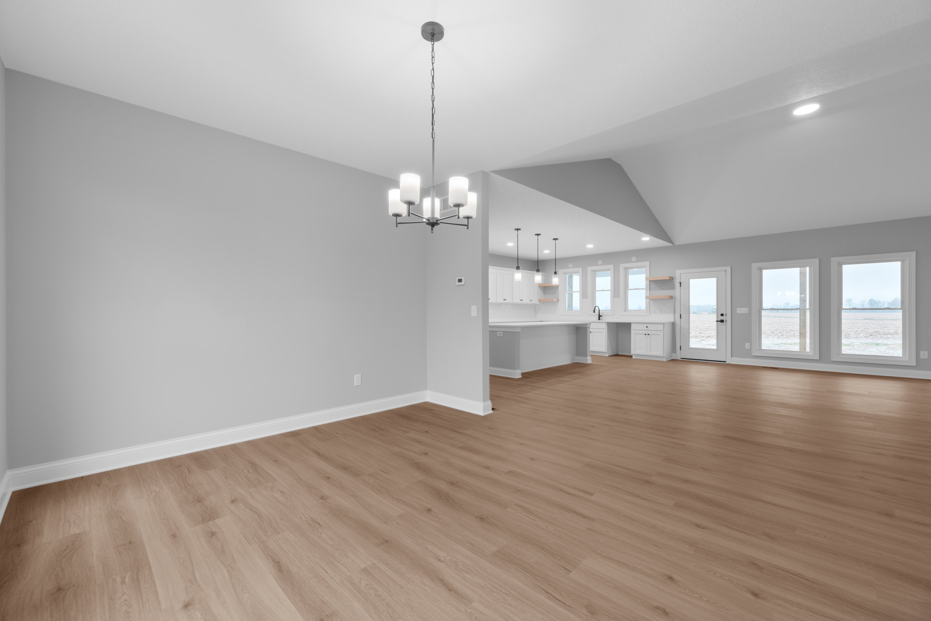 Open room with hardwood flooring, white plaster walls, large window and door overlooking a field and trees, modern chandelier hanging from ceiling.
