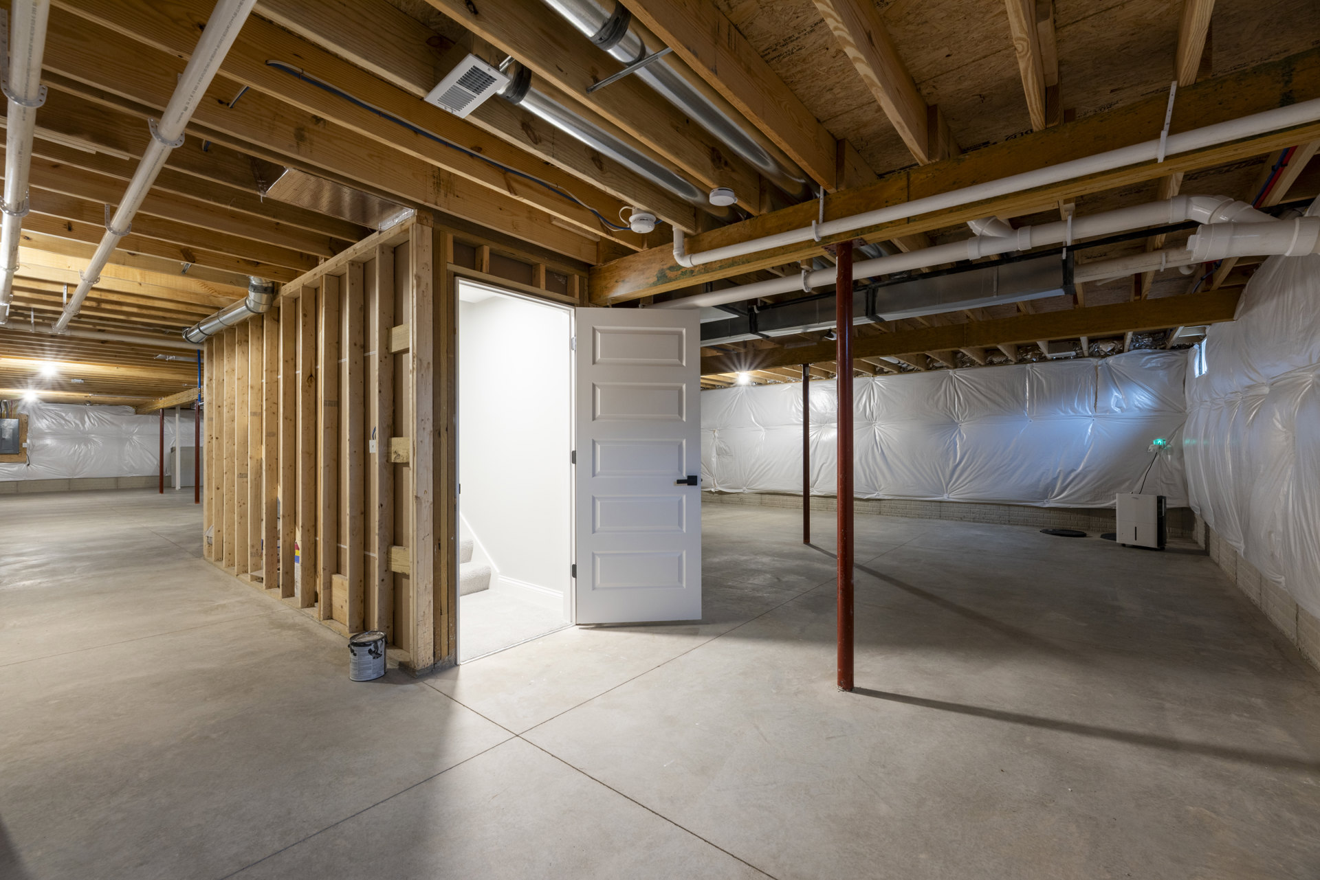 Open white door with black handle leading into a room featuring a wood accent wall, exposed metal pipes on wood ceiling, concrete floor, and plaster walls.