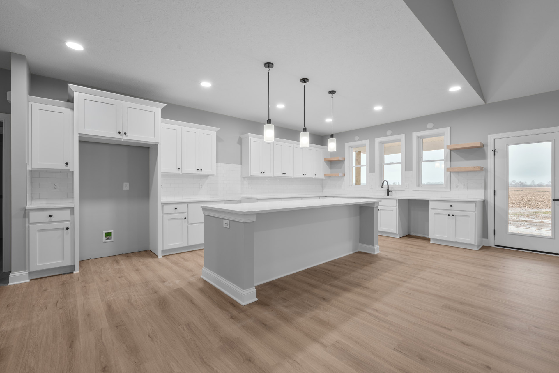 White kitchen with wood flooring, white cabinets, white countertops with column details, white walls, square-framed white door open to a field, window with white trim.
