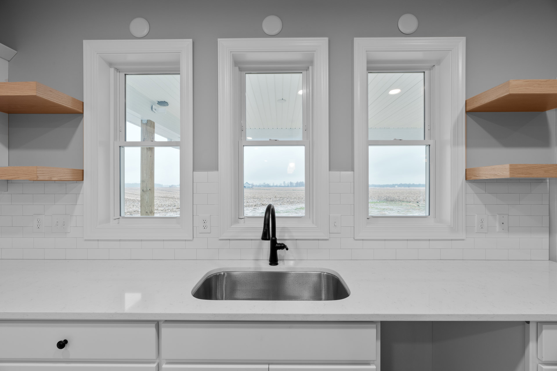 White kitchen with sleek countertops and cabinets, stainless steel sink, black faucet, large window overlooking a field, gray surface with white circle, soft lighting on window.