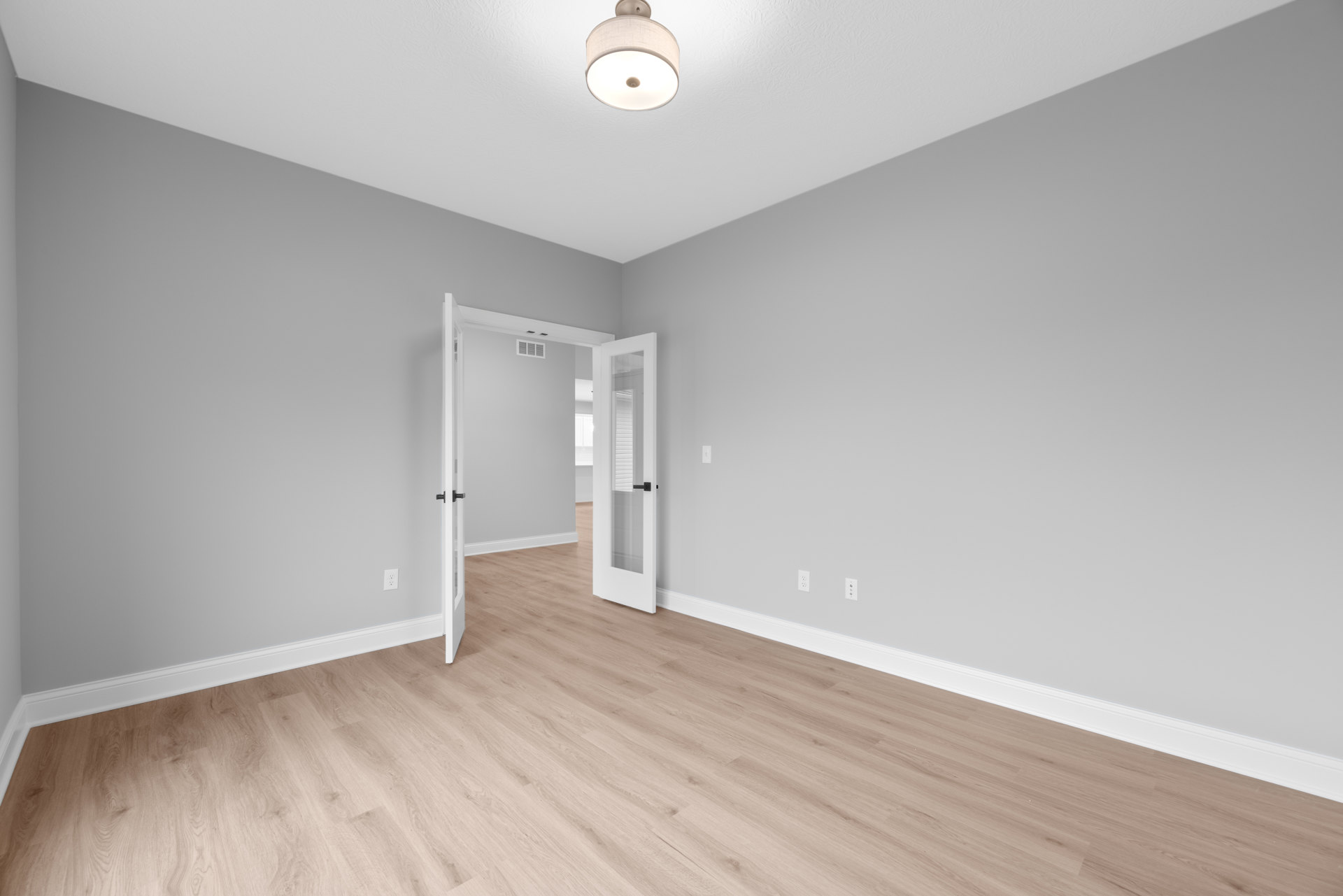 White paneled door with black handle opening into a room with light wood flooring, smooth white plaster walls, and a modern ceiling light fixture
