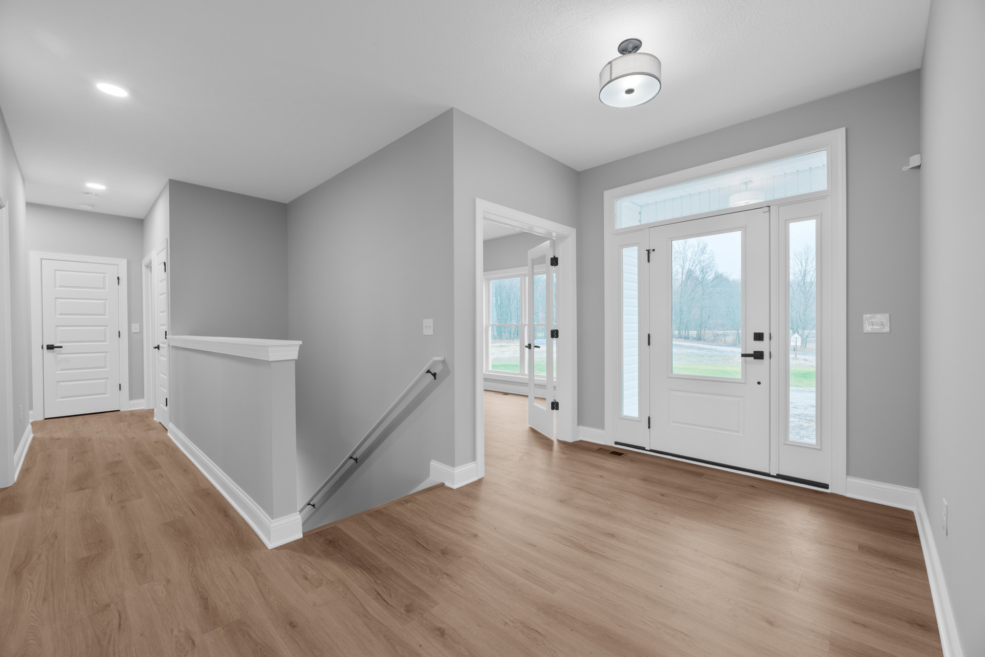 Hallway with light wood flooring, smooth white walls, white doors with black handles and glass panels, white ceiling, and modern light fixture with white shade
