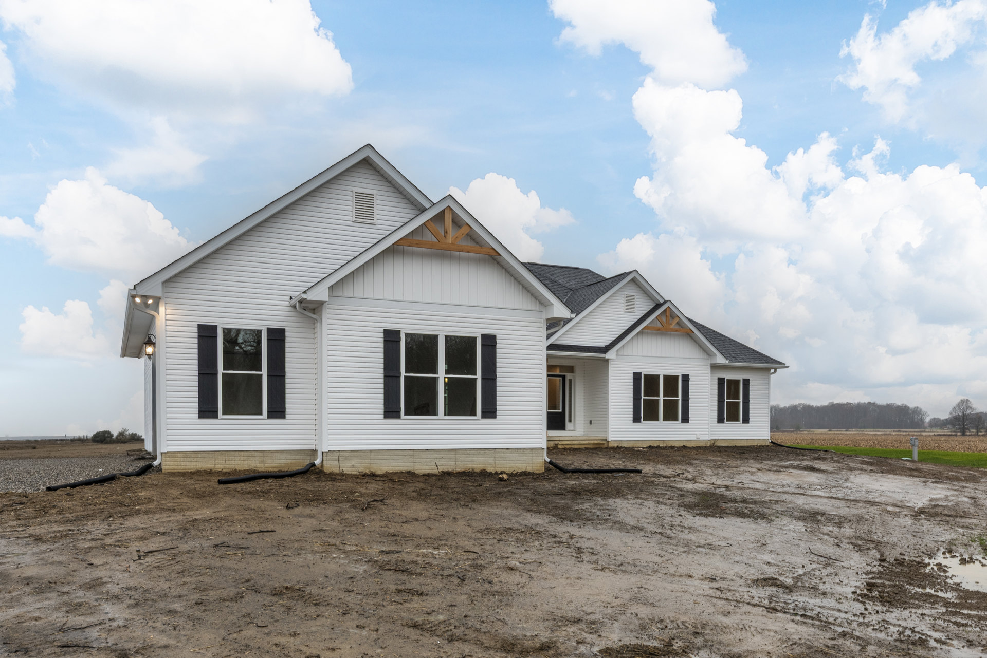 White house with black shutters and siding, dirt construction area in foreground, exposed pipe, cloudy blue sky overhead, illuminated window on front facade