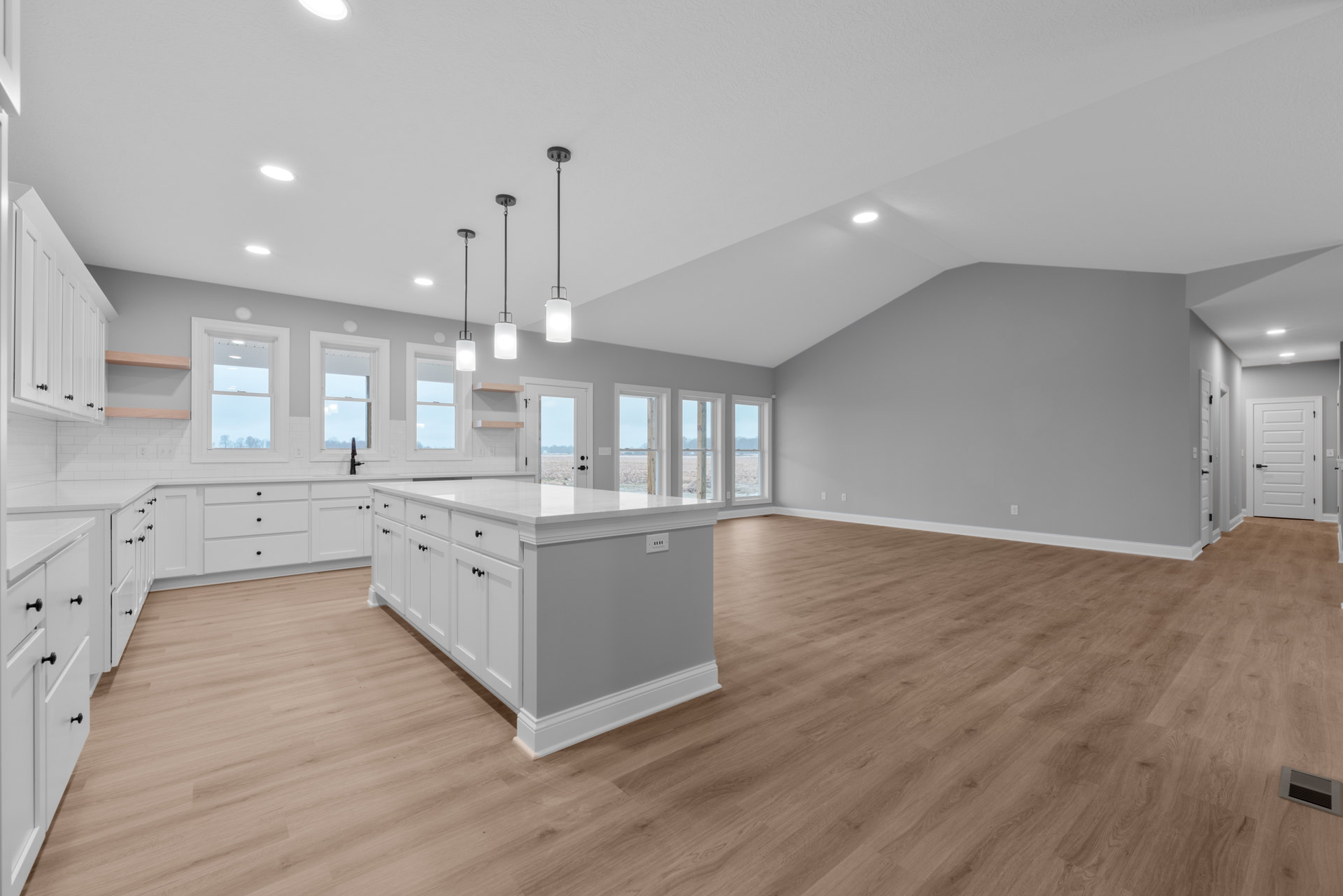 Open-concept kitchen with central island, hardwood floors, white cabinetry, stainless steel appliances, white door with black handles, large windows framed in white showing blue