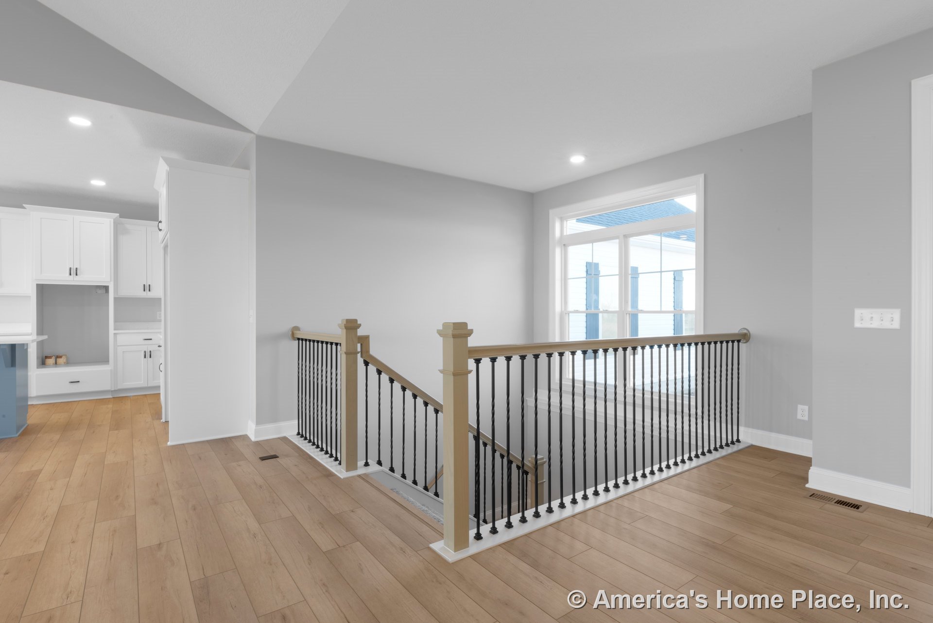 Stair landing with light wood plank flooring, black metal balusters on wood railing, large window with white trim, recessed ceiling lights, and adjacent open kitchen featuring