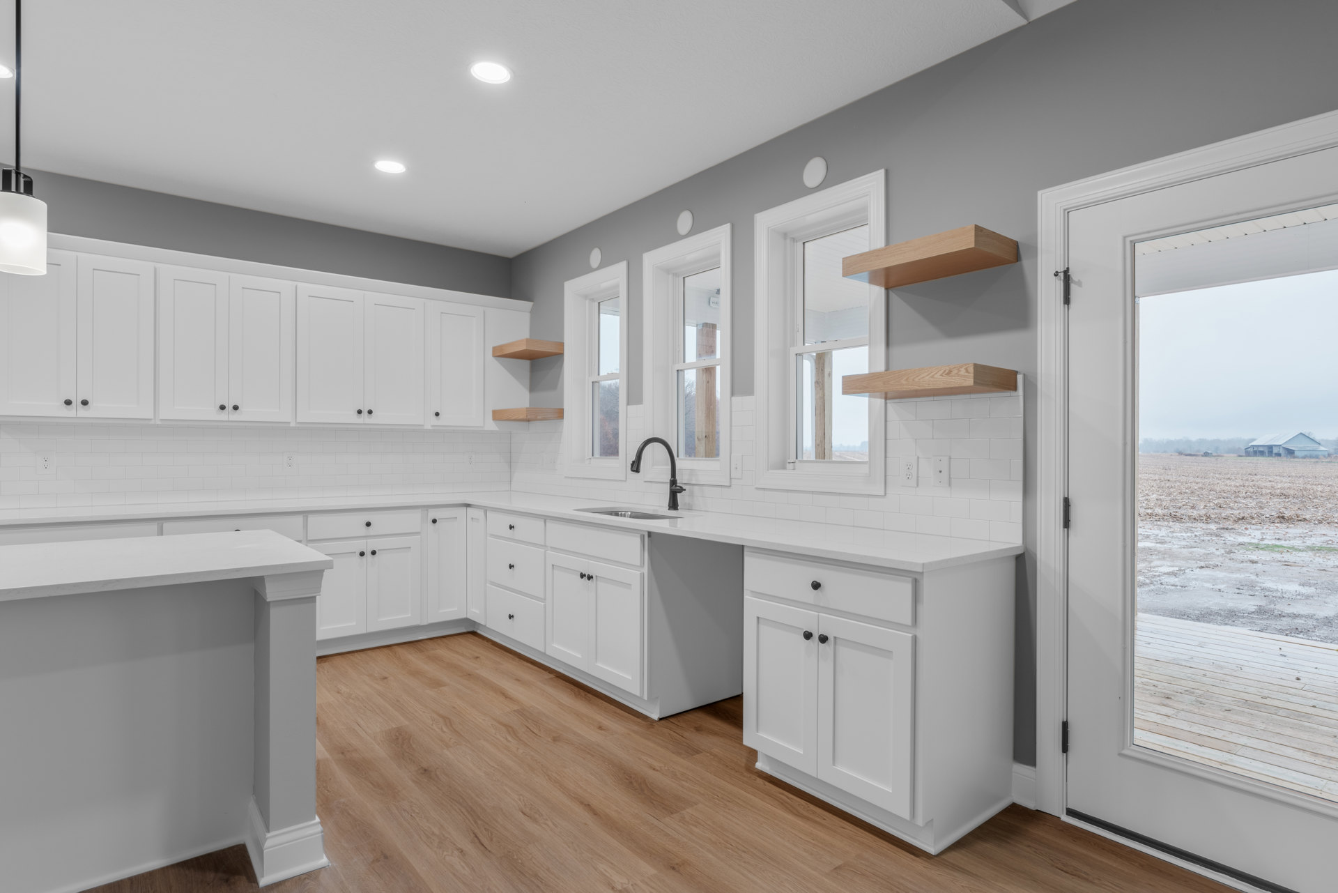 White kitchen cabinets with black hardware, wood plank flooring, white quartz countertop, undermount sink with black faucet, open doorway leading to outdoor farm view, white