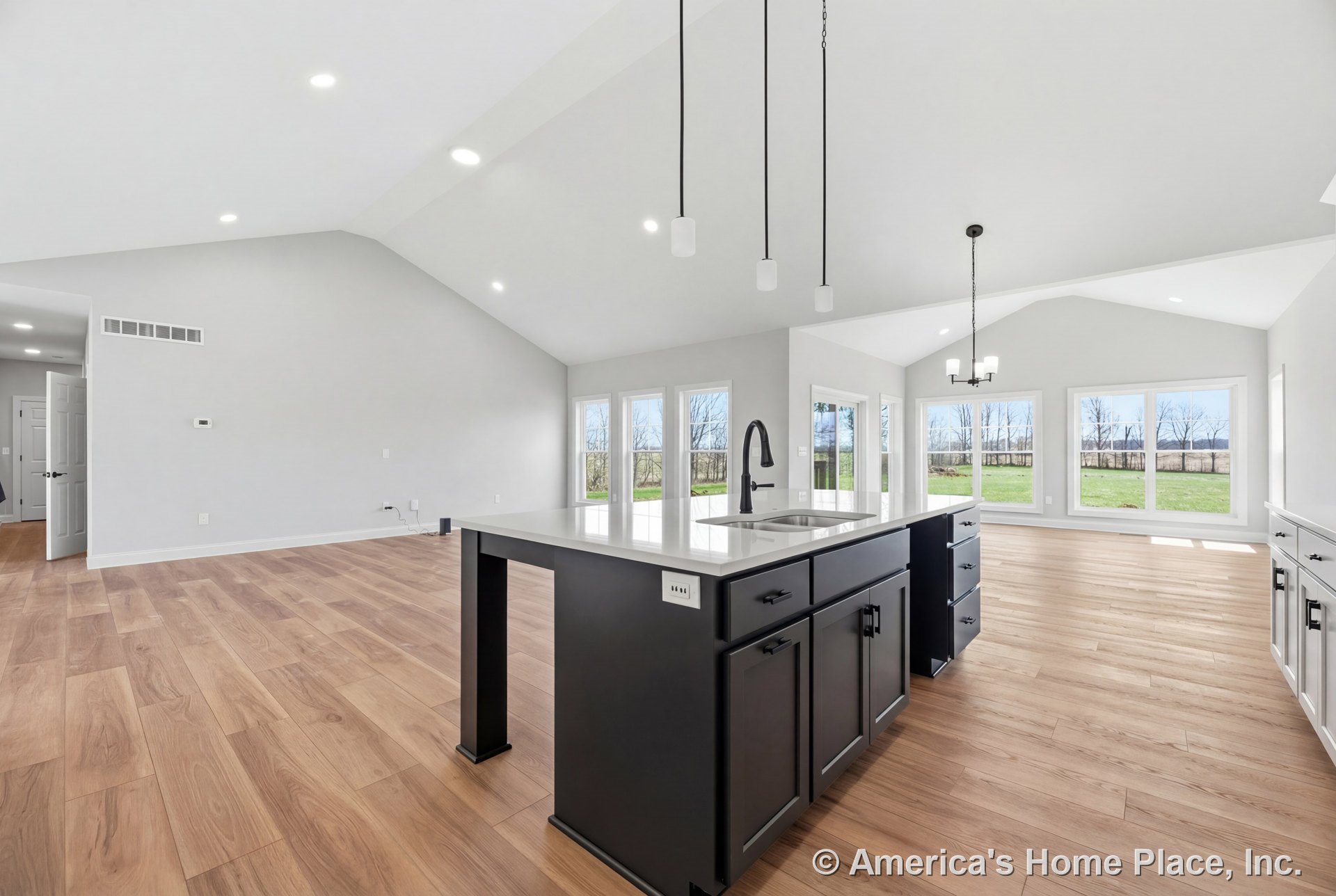 Vaulted ceiling with recessed lighting above open kitchen and living area, large windows along back wall, central kitchen island featuring dark cabinetry, quartz countertop